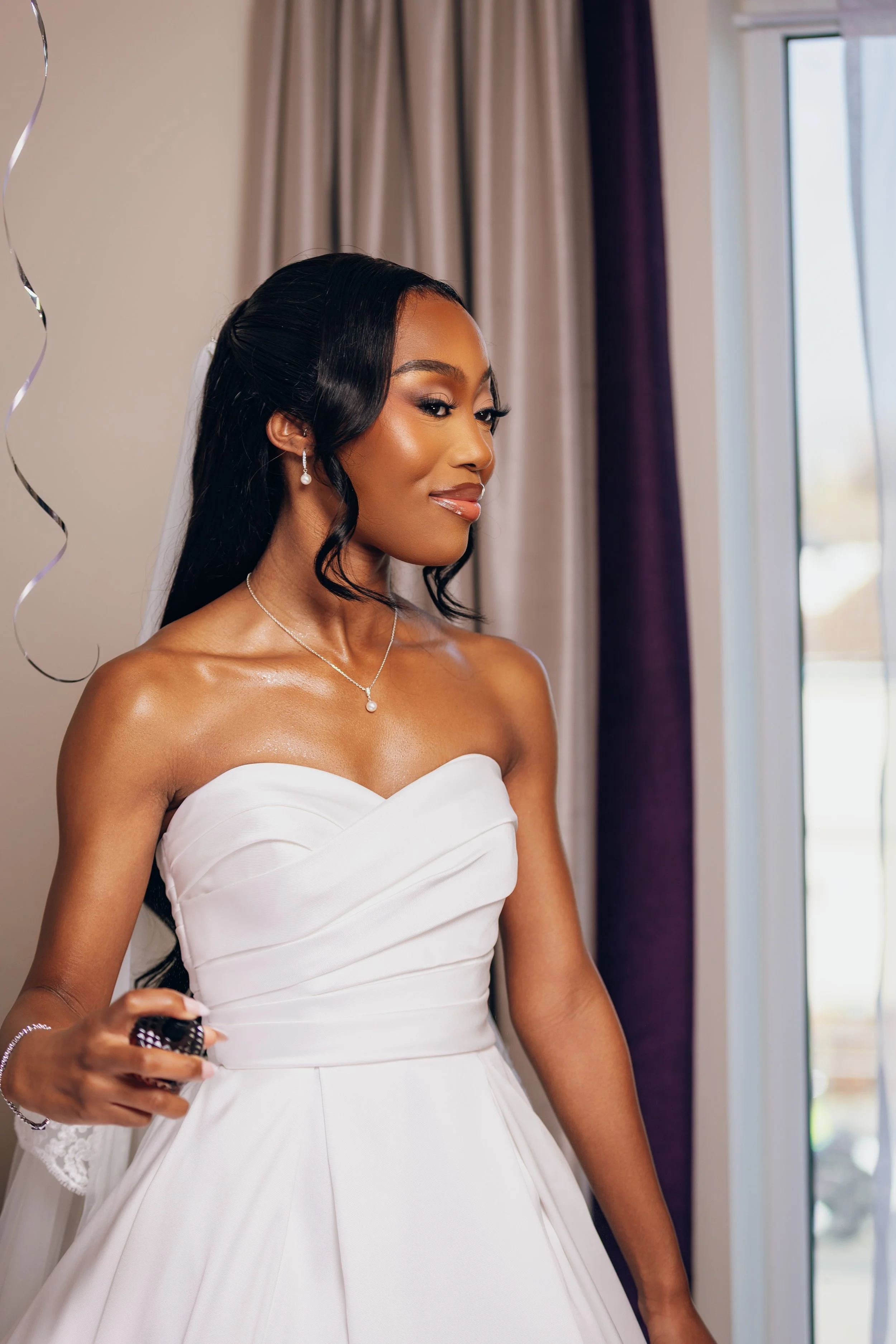 A bride in a white strapless wedding dress with dark, wavy hair and pearl jewelry, standing indoors near a window with curtains in the background.