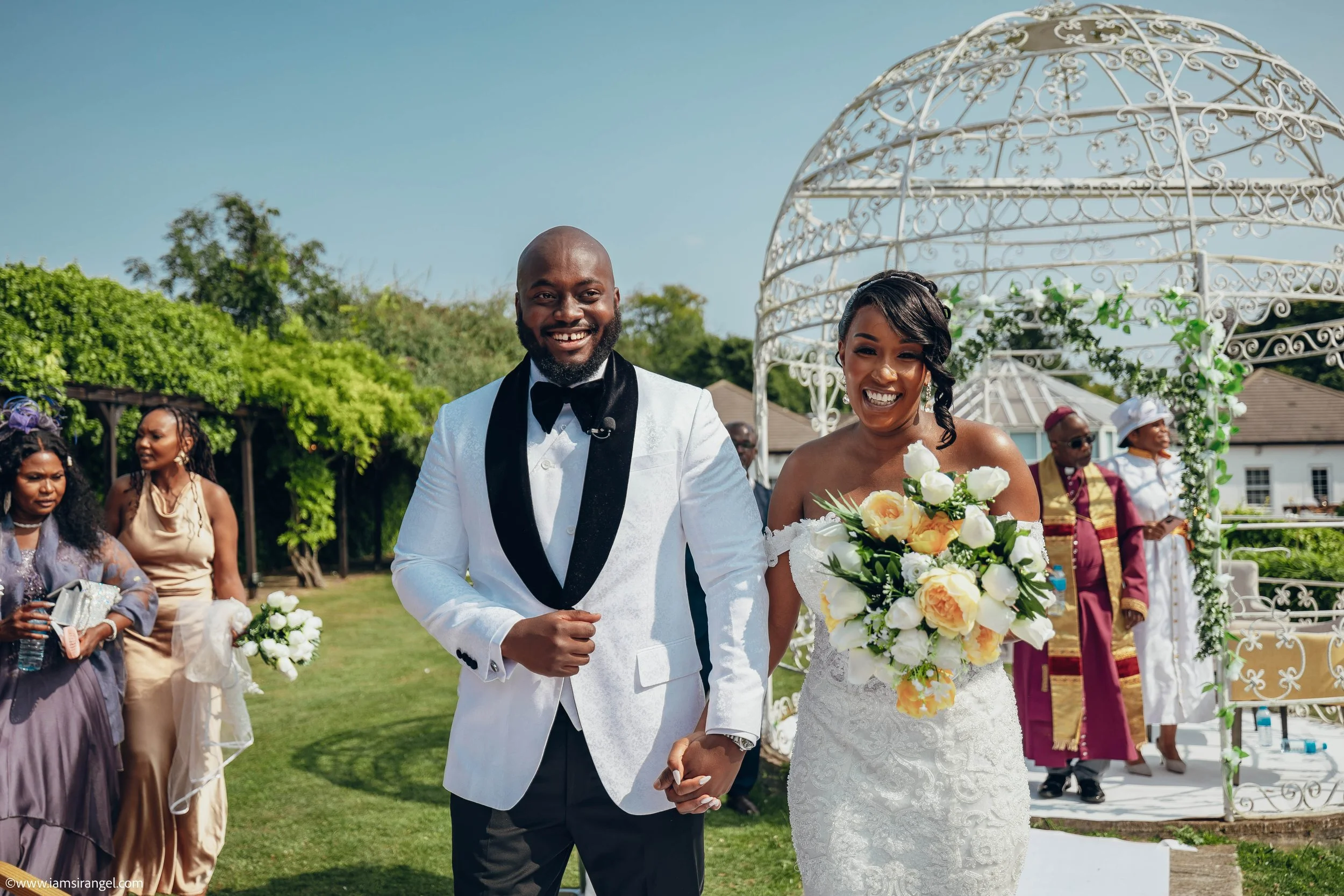 A happy bride and groom walking hand in hand at their outdoor wedding ceremony, surrounded by friends and family, with a white ornate gazebo and green trees in the background.