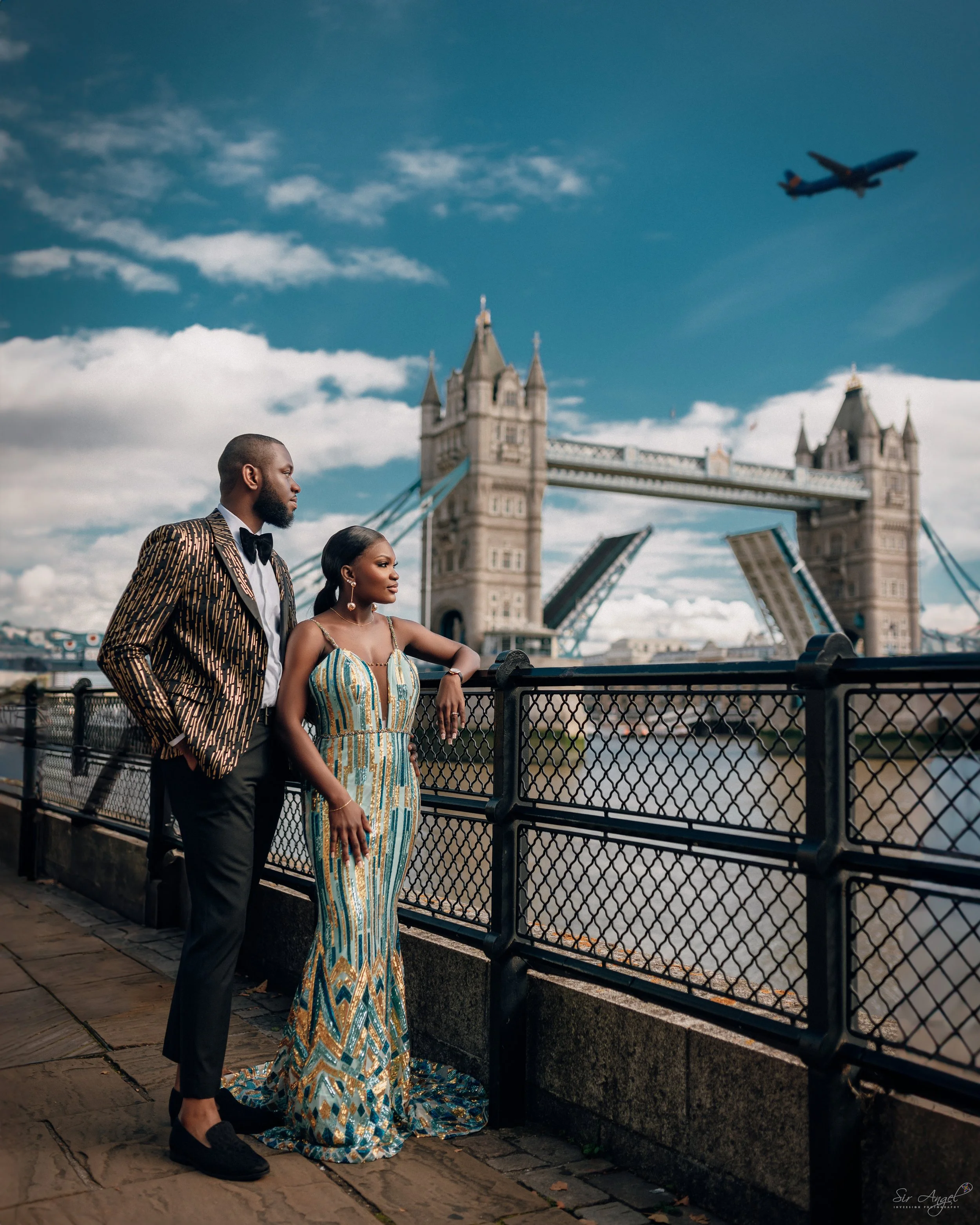A couple dressed in elegant attire standing by the Thames River near Tower Bridge in London, with an airplane flying overhead in the blue sky.
