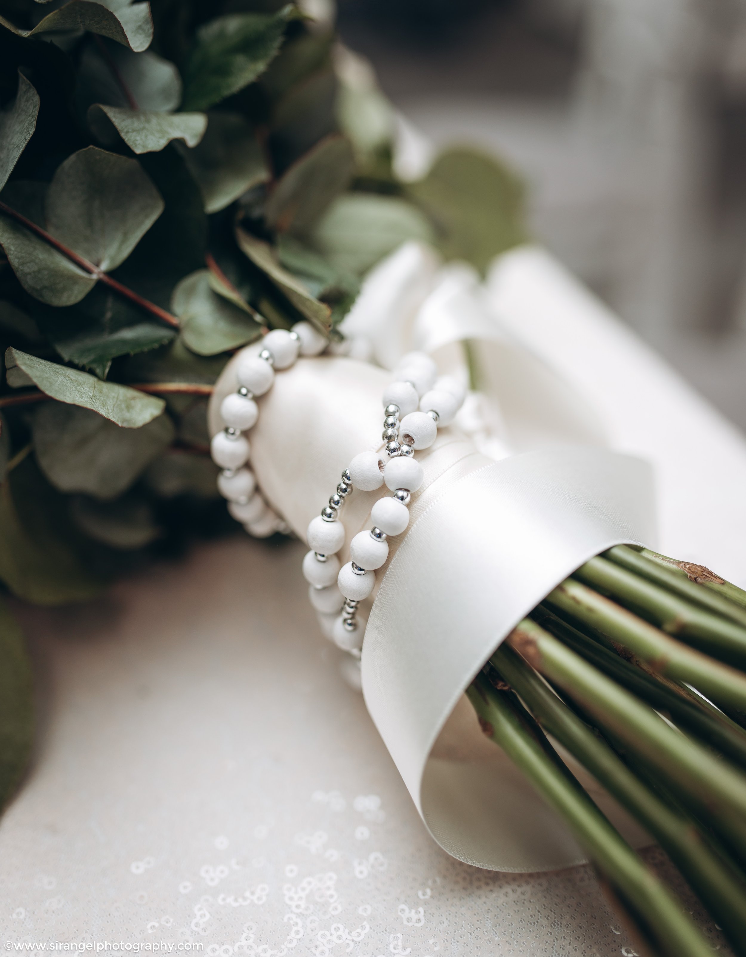 Close-up of a bridal bouquet with greenery, white ribbon, and pearl and silver bead accents.