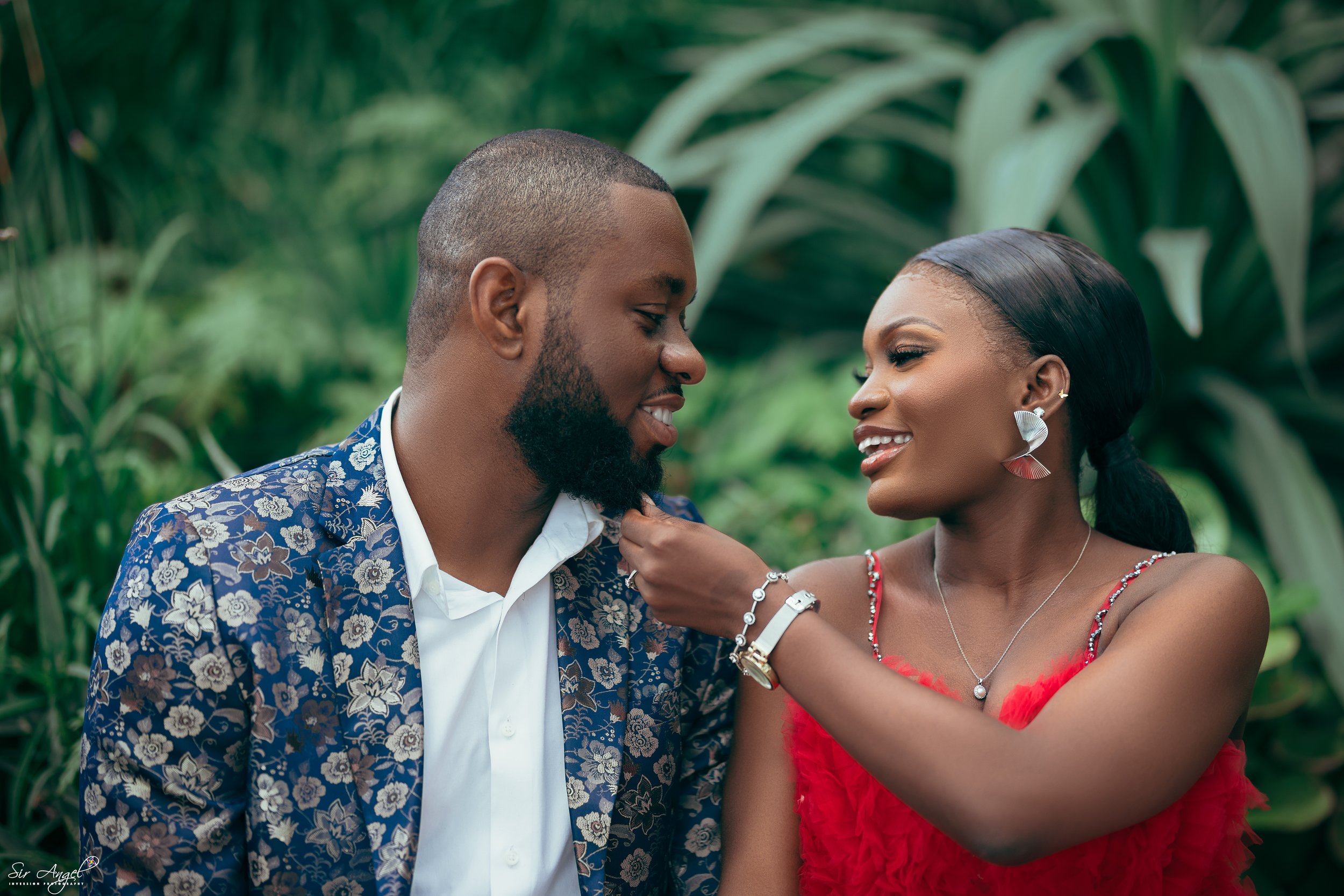 A romantic moment between a man and woman outdoors, with the woman touching the man's face, both smiling, surrounded by lush green plants.