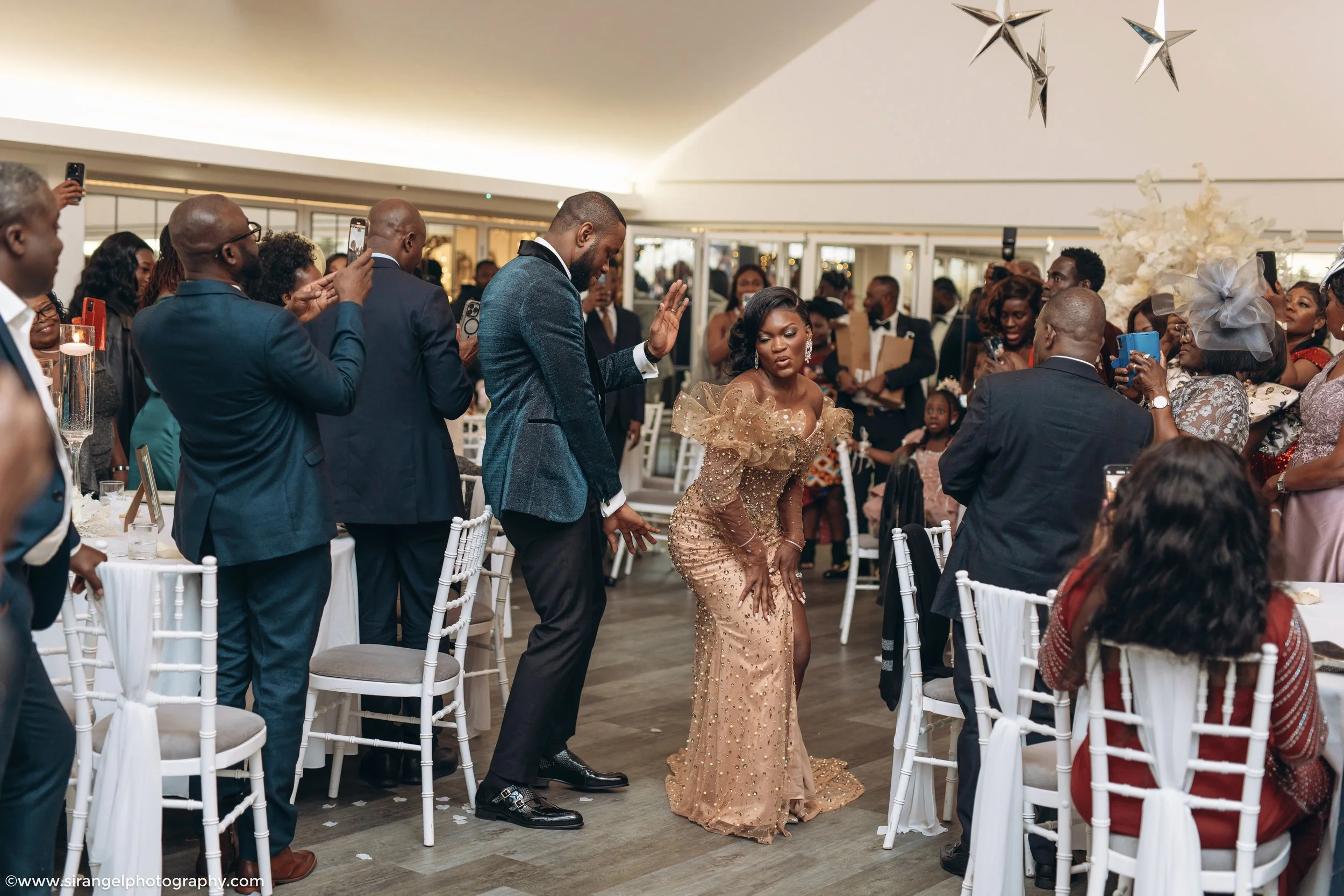A woman in a glamorous gold dress dancing at a formal event surrounded by seated and standing guests taking photos and videos.