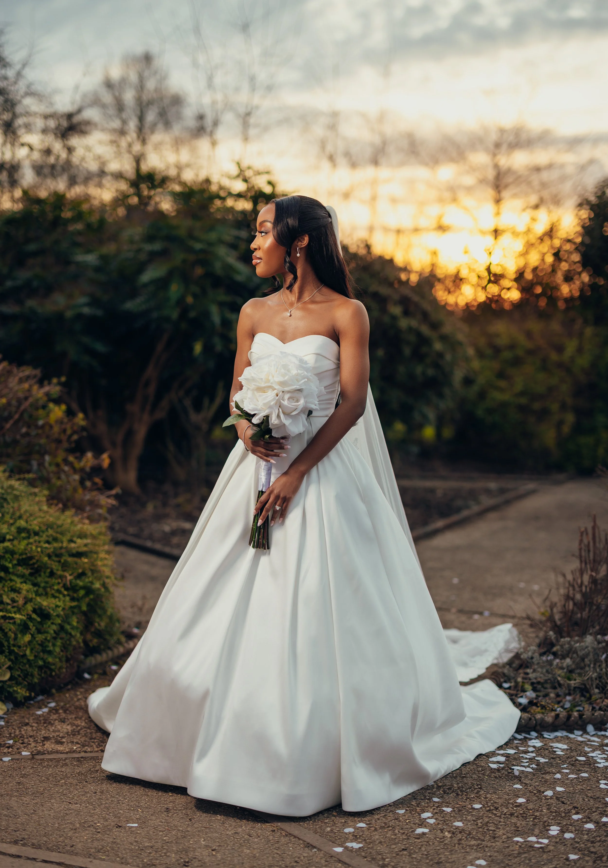 A bride in a strapless white wedding gown holding a bouquet of large white flowers, standing outdoors at sunset with trees and bushes in the background.
