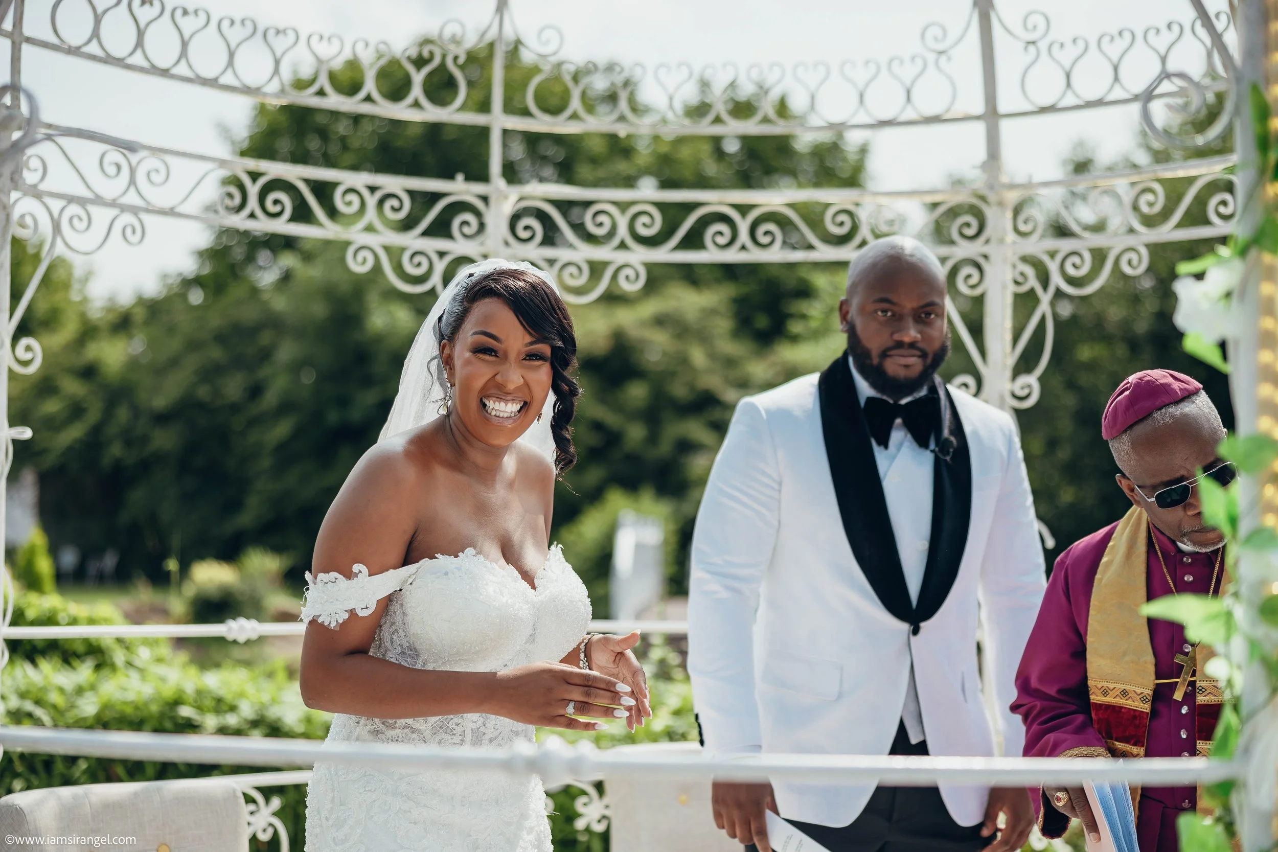 A bride in a white wedding dress smiling on her wedding day, standing next to a groom in a white tuxedo with a black lapel, outdoors during the day.