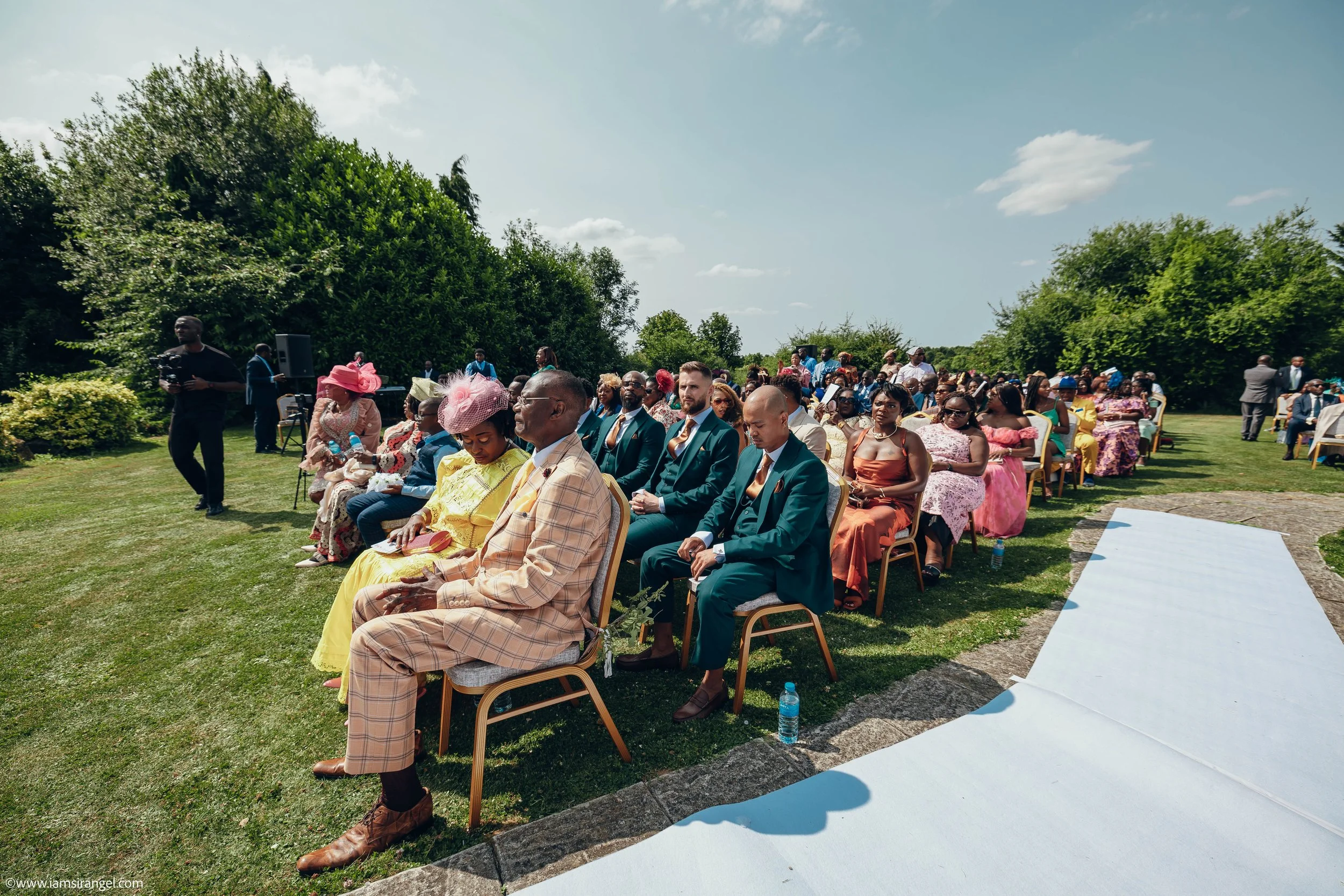 Outdoor wedding ceremony with guests seated on chairs, dressed in colorful and formal attire, under a partly cloudy sky surrounded by greenery.