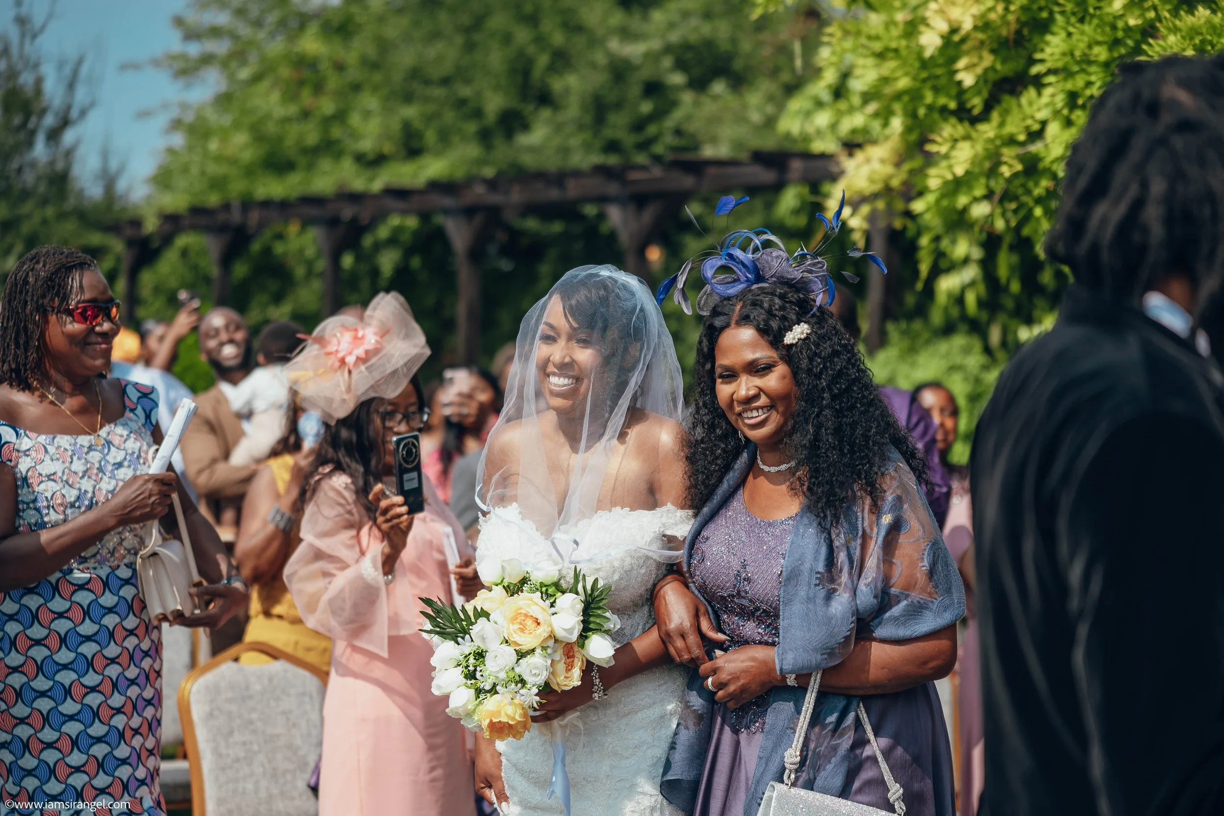 A bride in a white wedding dress with a veil and holding a bouquet of white and yellow flowers, smiling and standing next to a woman in a purple dress with a purple fascinator hat, at an outdoor wedding ceremony. Guests are in the background, some ta