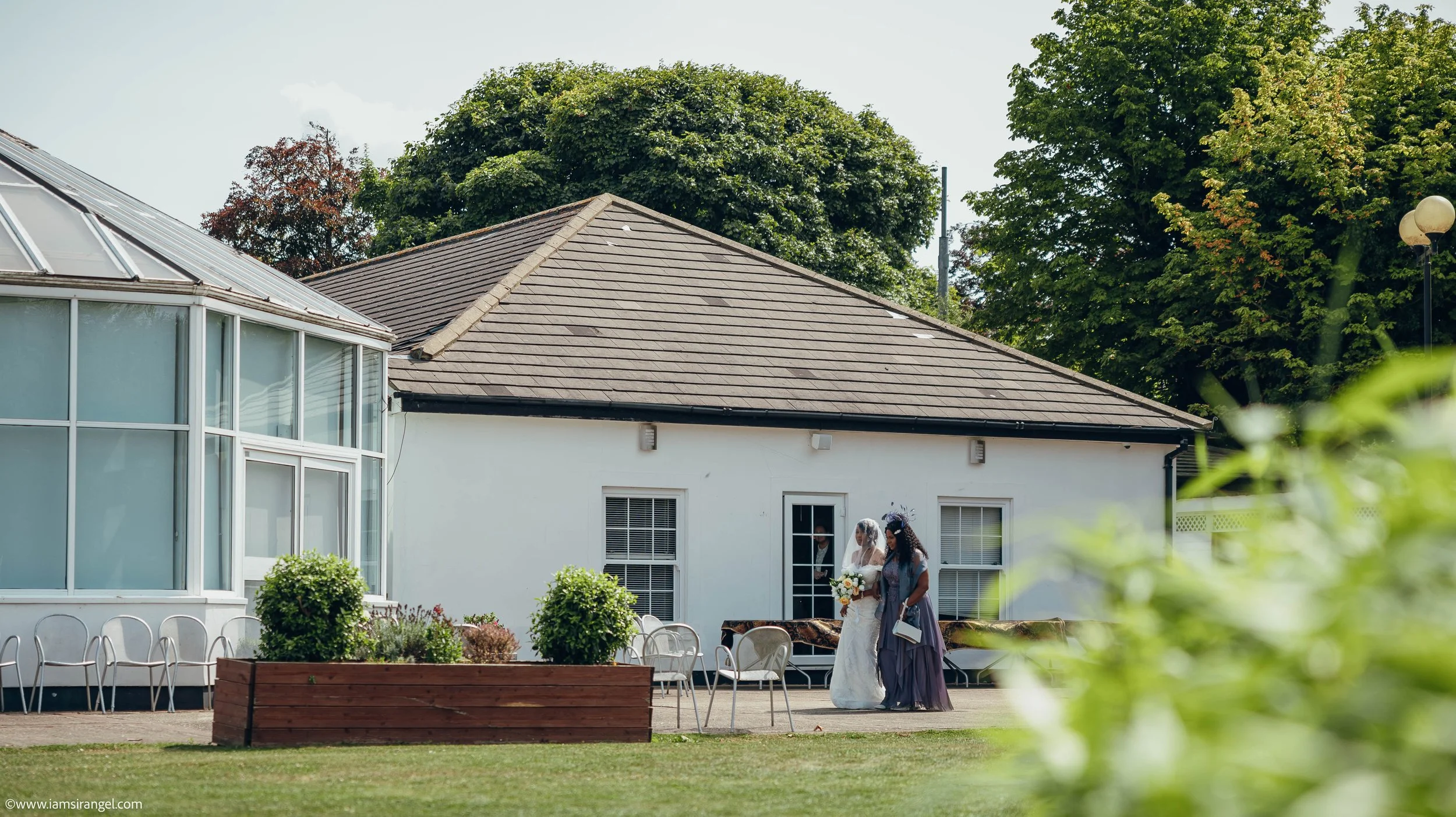 A bride in a white wedding dress and veil holding a bouquet, walking with a bridesmaid in a dark gown in an outdoor area near a white building with a brown roof, surrounded by green trees and chairs.