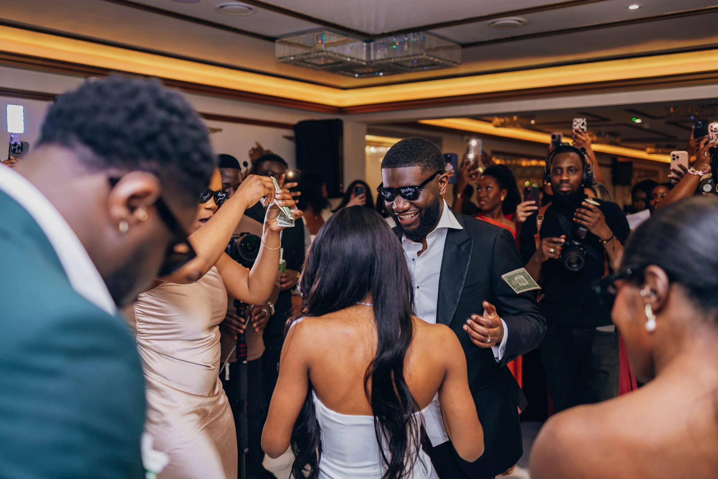 A wedding reception with a bride and groom dancing among guests, some of whom are taking photos. The groom is wearing sunglasses and a suit with dollar bills attached to his clothing. The bride has long dark hair and is wearing a strapless white wedd