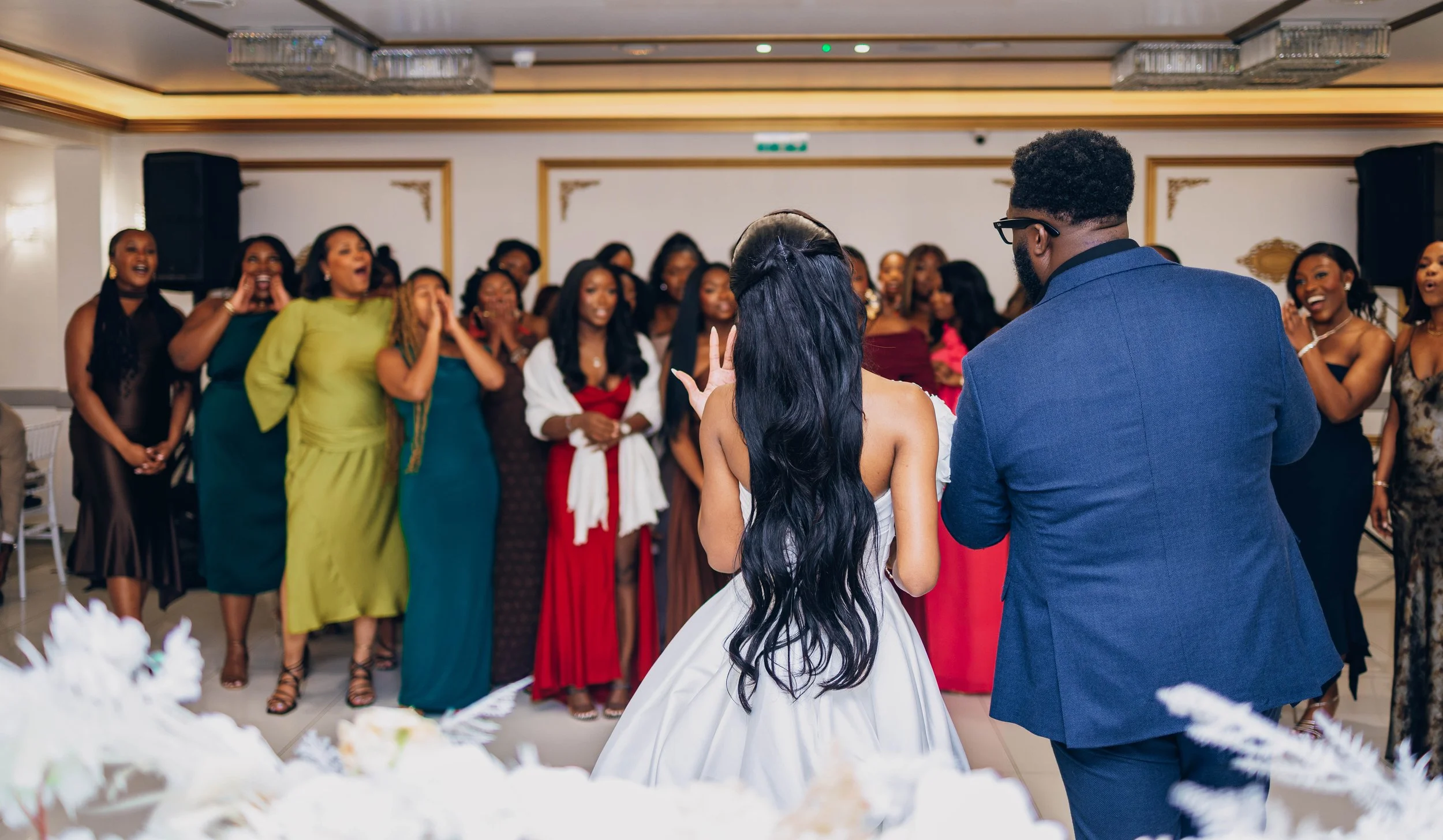 Couple dancing at a wedding reception surrounded by women in formal dresses, in an elegant ballroom with chandeliers.
