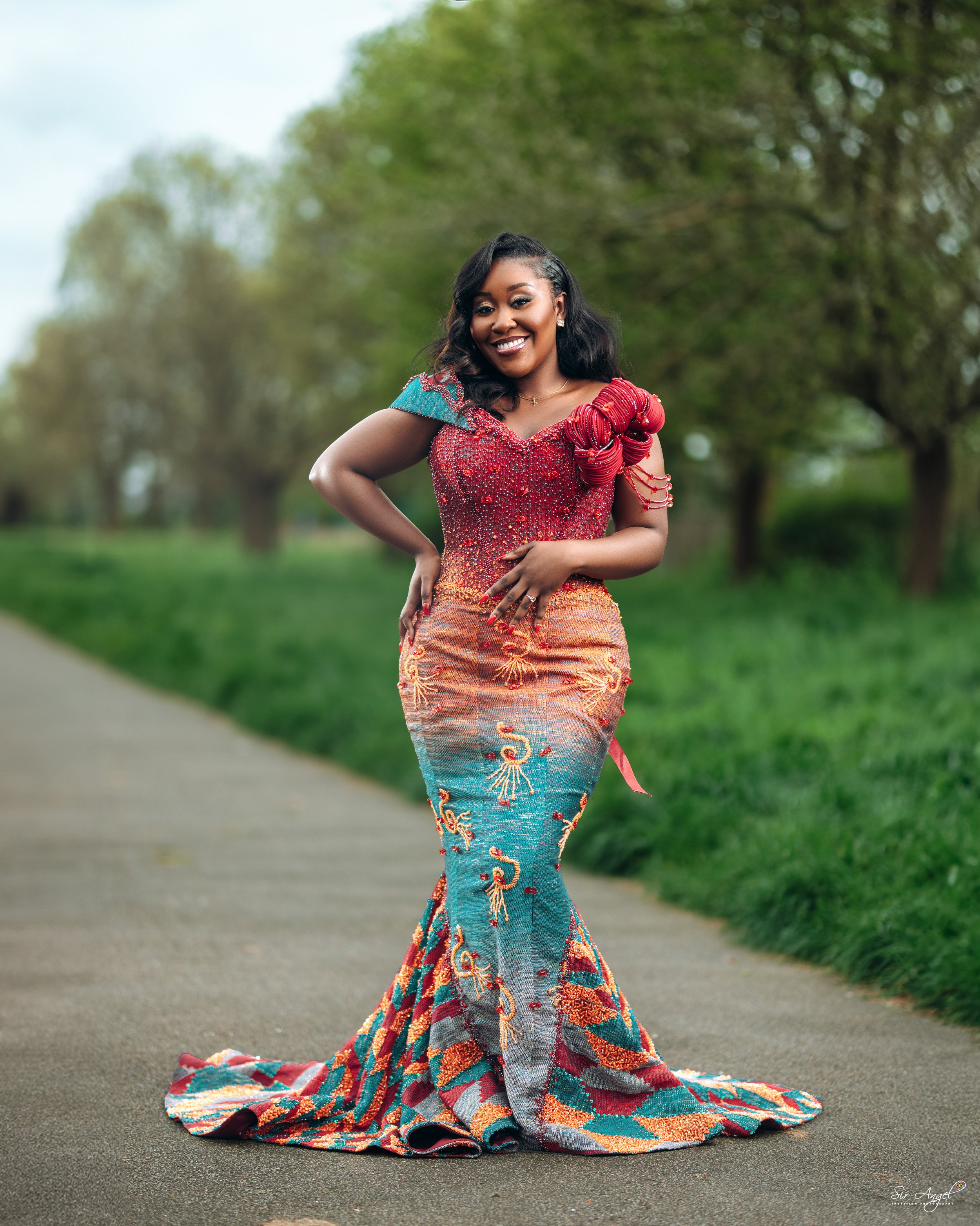A woman wearing a colorful, elaborate dress with embroidery stands on a pathway in a park with green grass and trees, smiling at the camera.