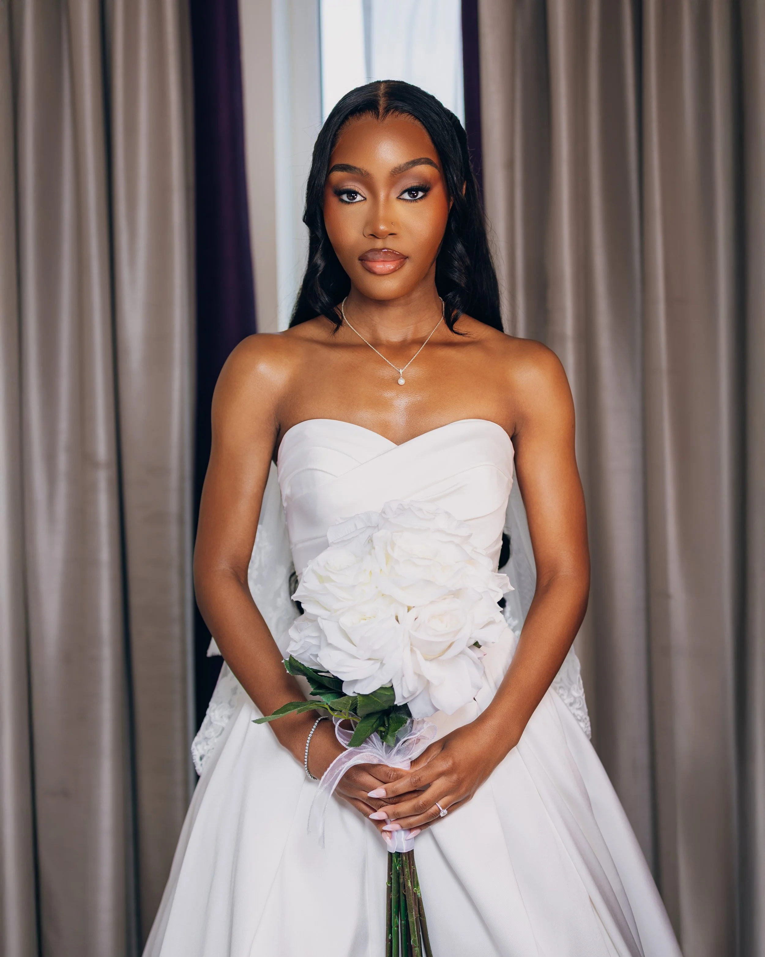 A woman in a white strapless wedding dress holding a bouquet of white roses, standing indoors with curtains in the background.