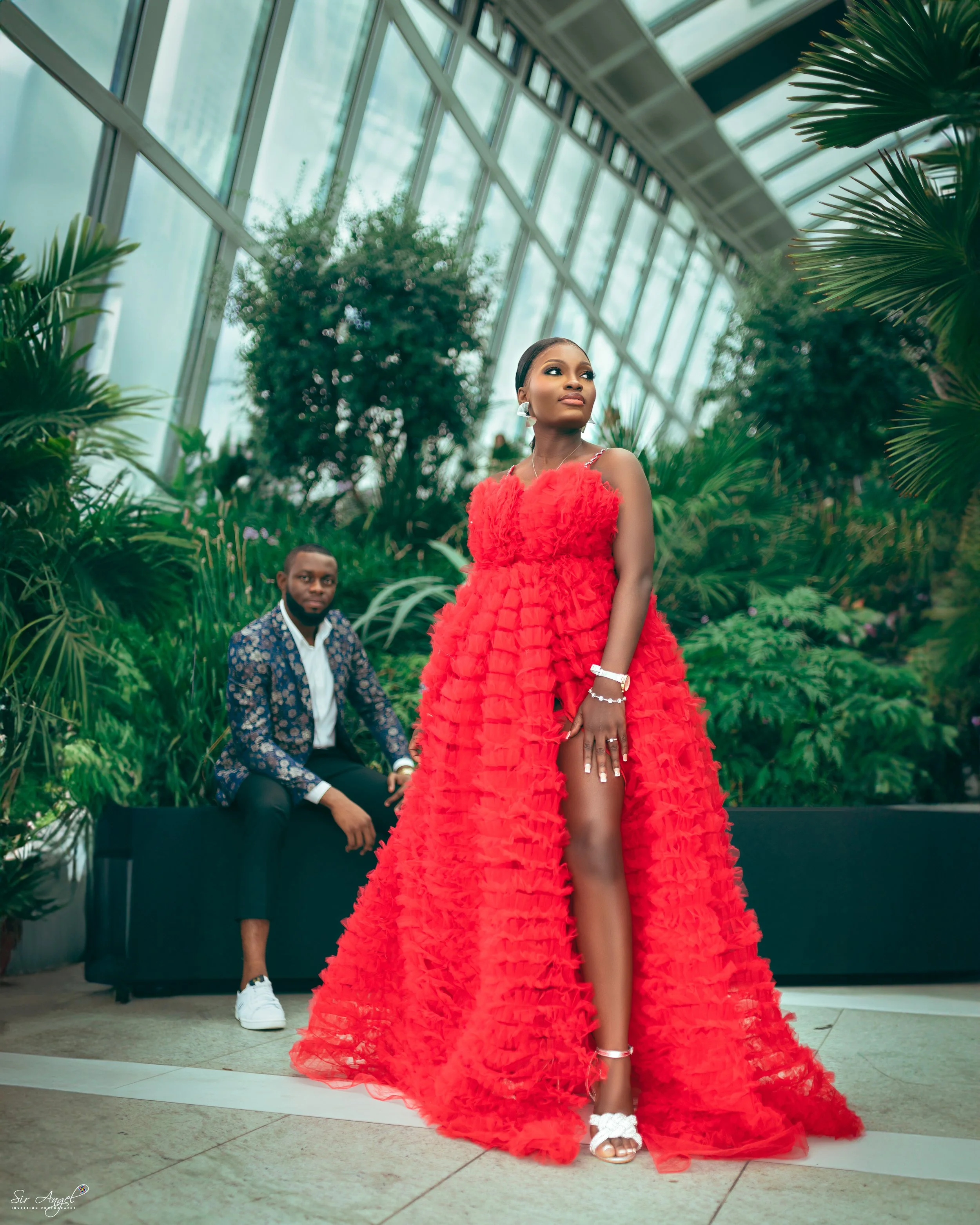 A woman in a vibrant red, ruffled evening gown with a high slit, standing in a modern greenhouse or botanical conservatory, with a man in the background seated on a black bench, surrounded by lush green plants and tall glass windows.
