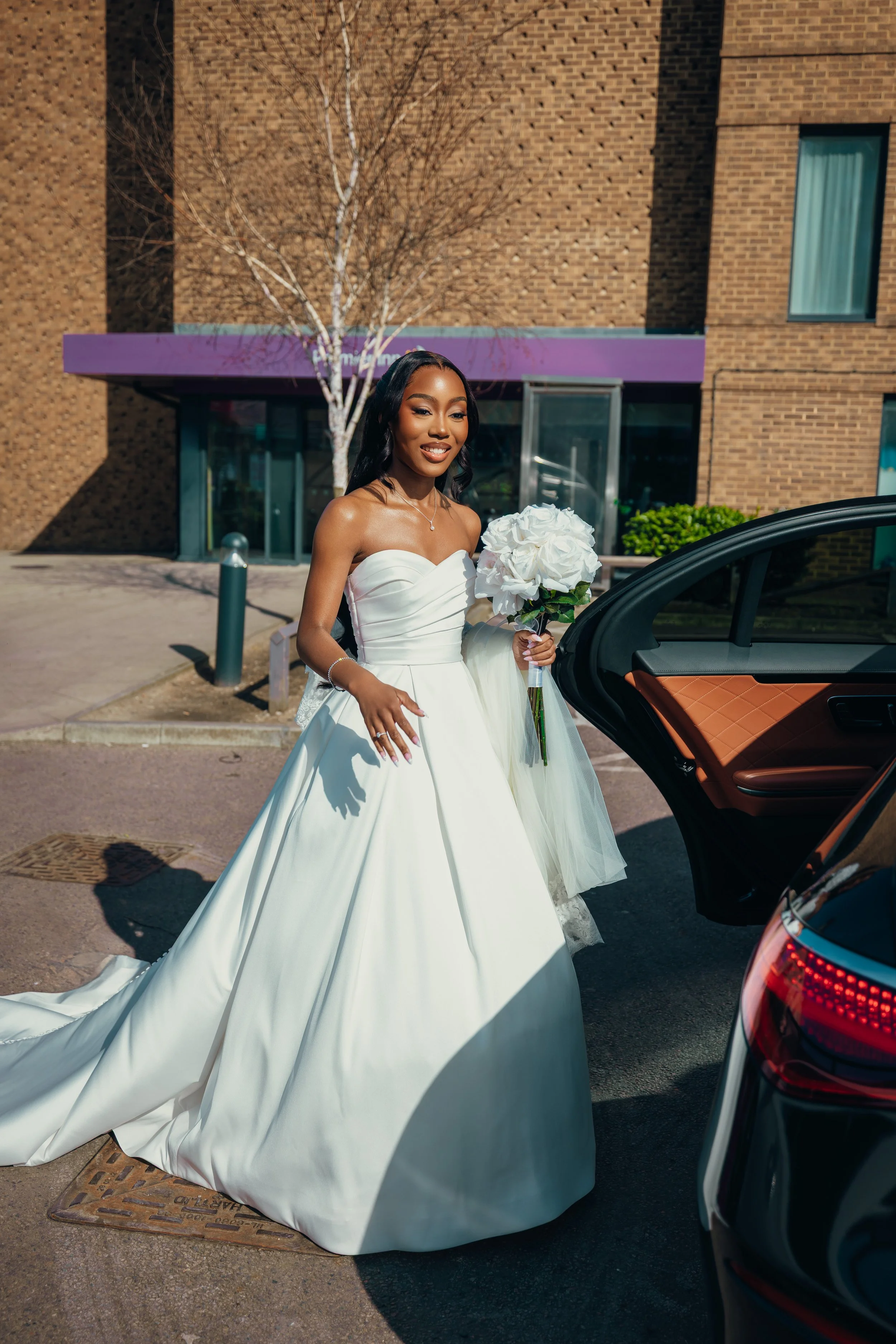 A bride in a white wedding gown holding a bouquet of white flowers outdoors next to a black car.