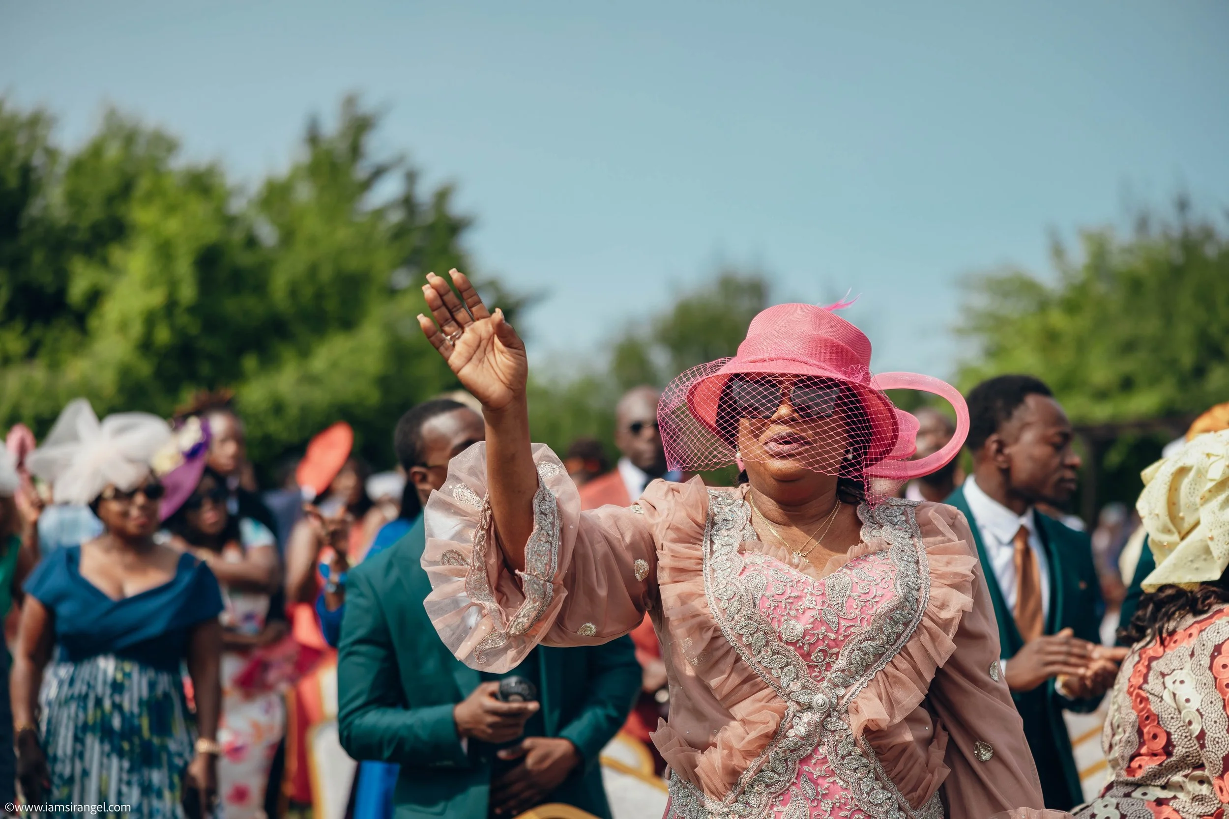 A woman in a pink dress and large pink hat with a veil is raising her hand at an outdoor event, surrounded by other attendees dressed in formal and colorful attire.