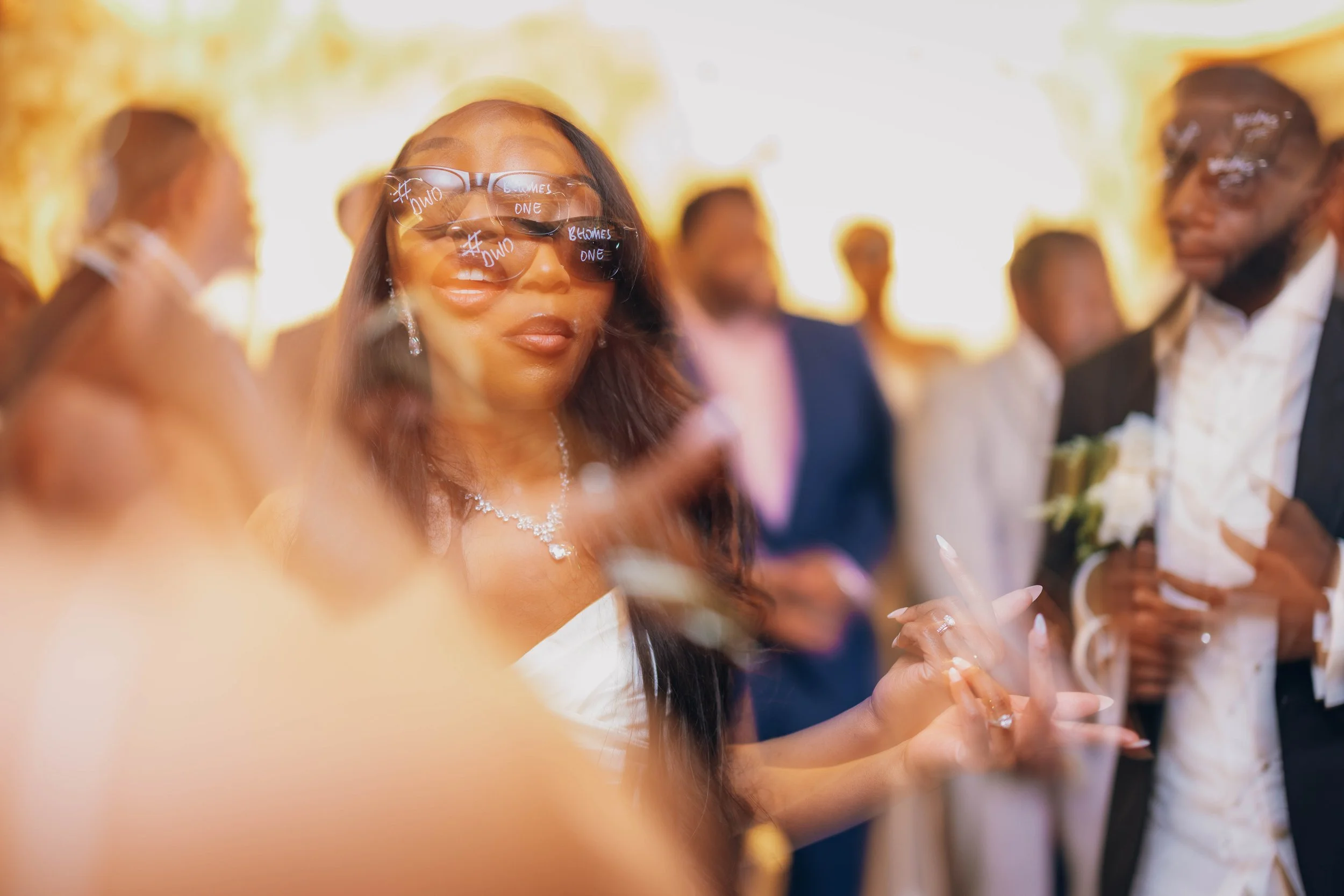 People at a wedding reception, woman in sunglasses and jewelry, man in suit holding a bouquet.