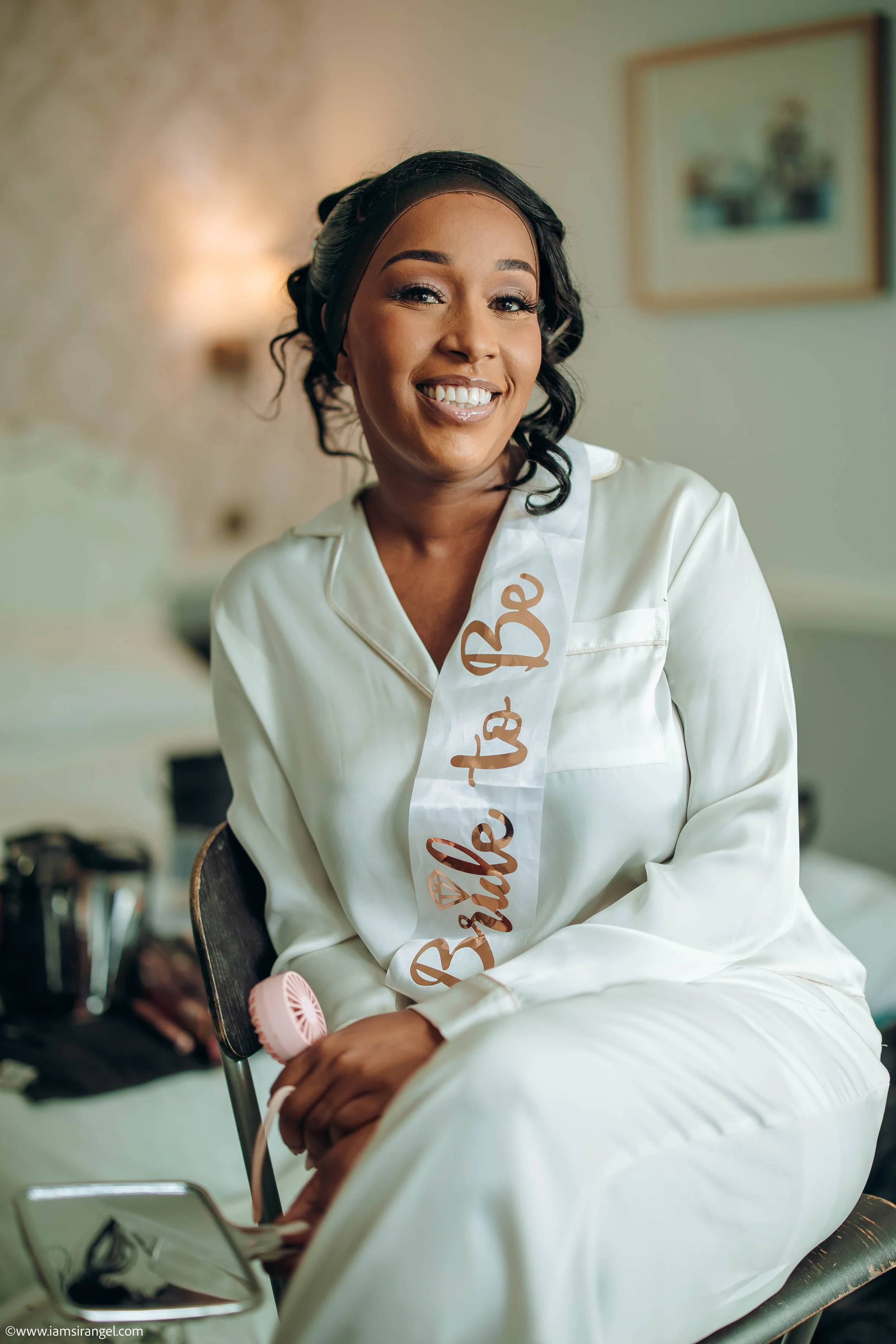 Woman in white satin pajamas sitting on a bed with a 'Bride to Be' sash, smiling and holding a small handheld fan.