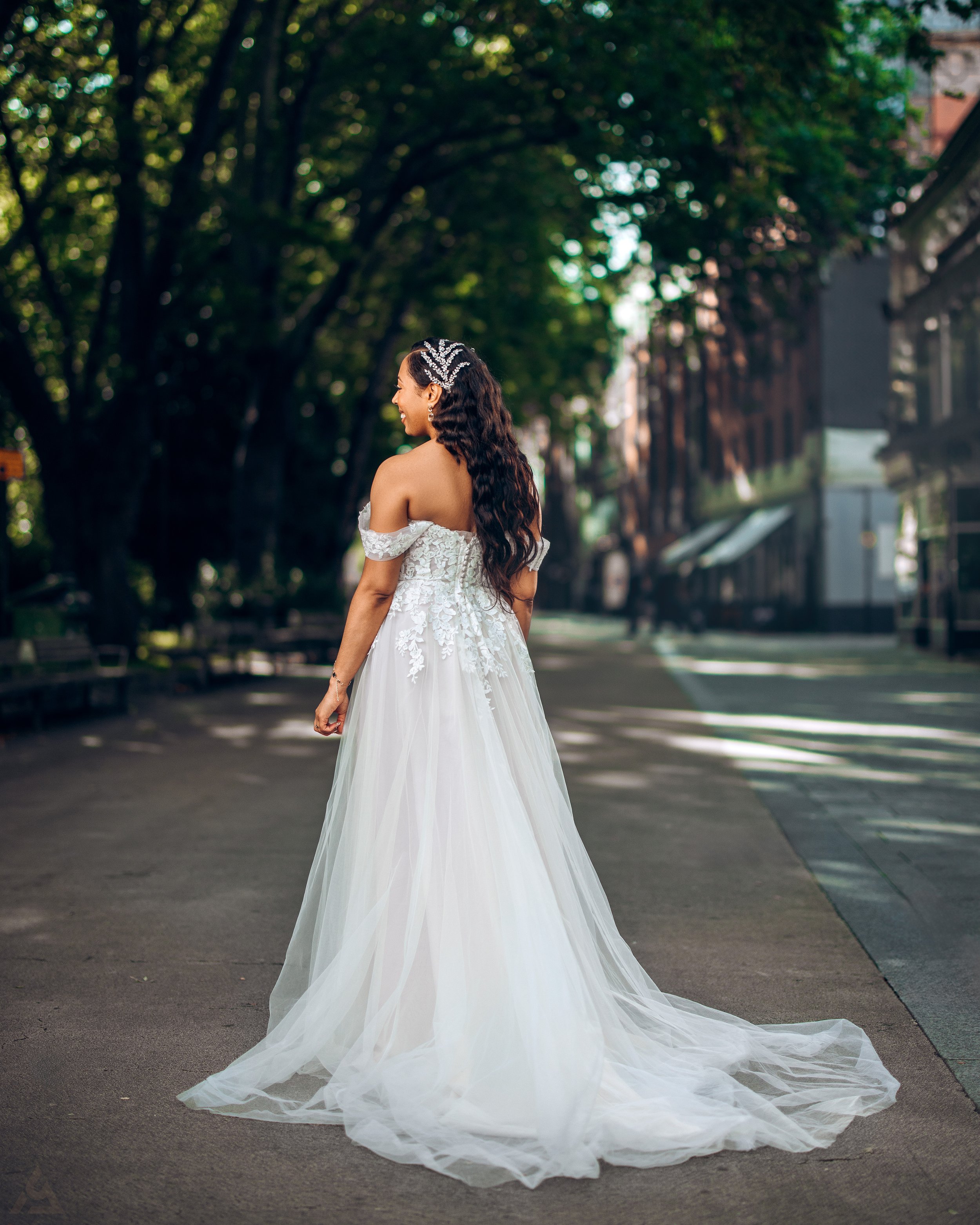 A woman in a white wedding dress walking on a city street lined with trees.