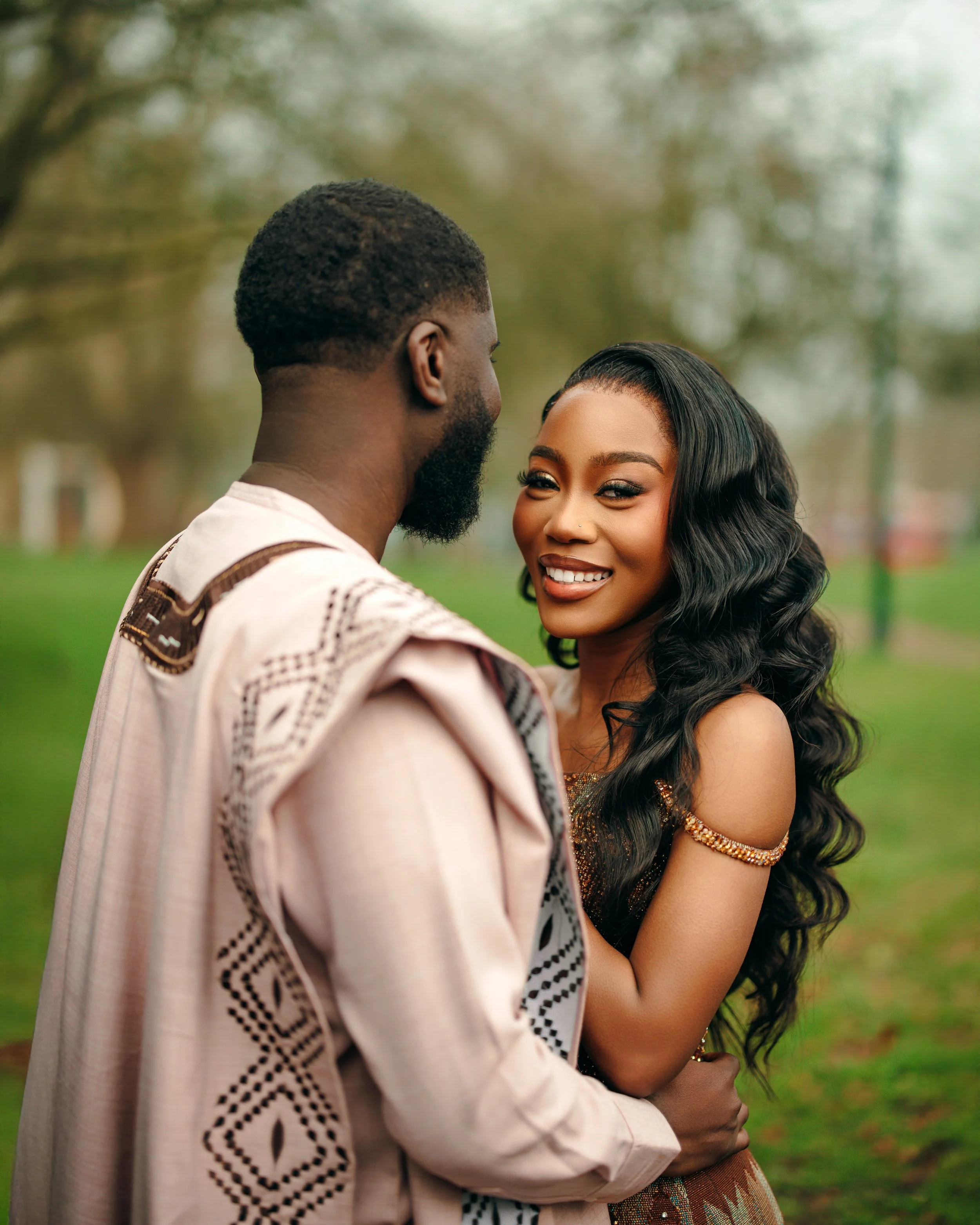 A smiling woman with long, curly black hair, wearing a sleeveless dress and gold jewelry, is embraced by a man with a beard and short curly hair, wearing a patterned traditional shirt. They are outdoors in a park with trees.