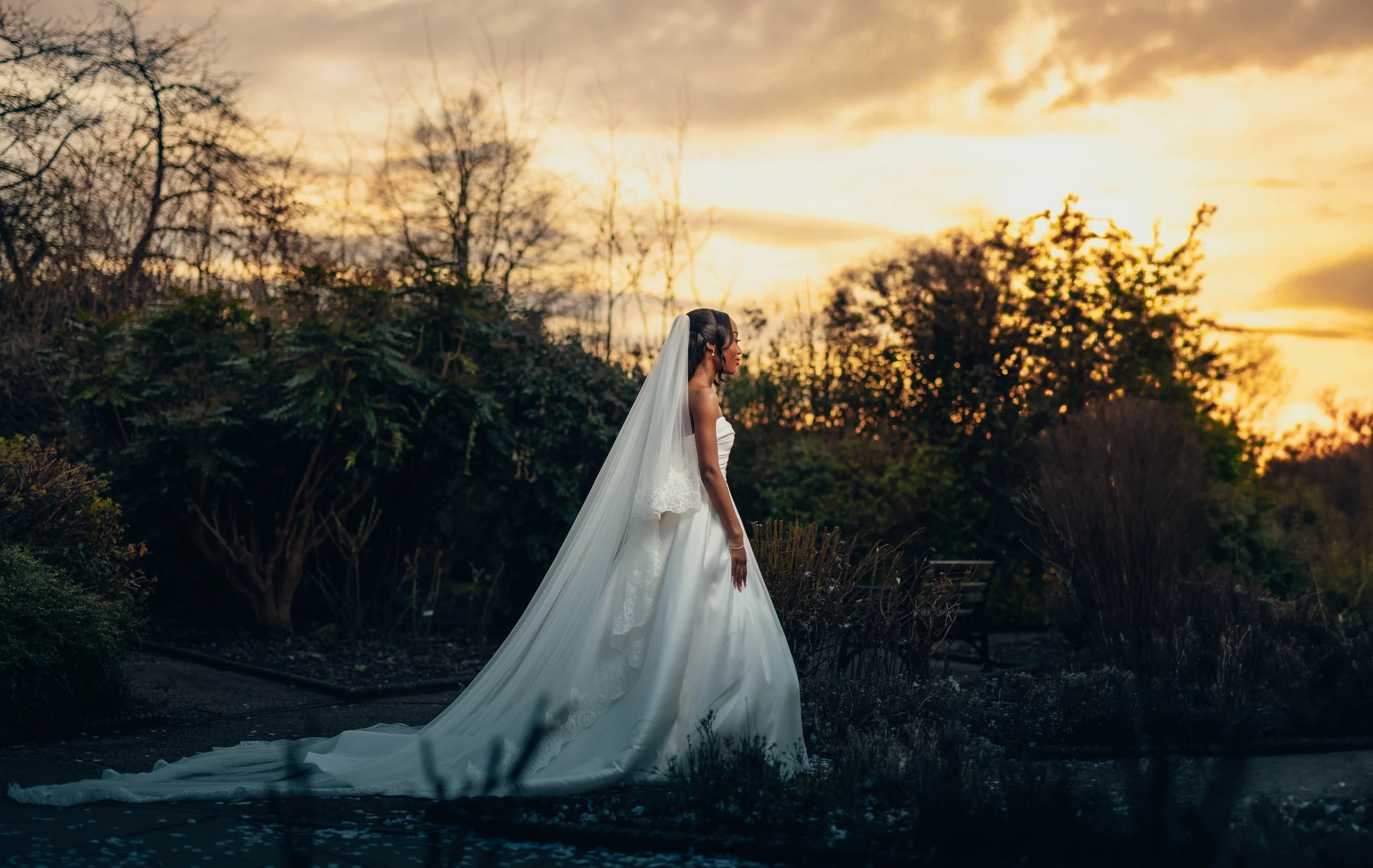 A bride in a white wedding dress with a long train and veil standing outdoors at sunset.
