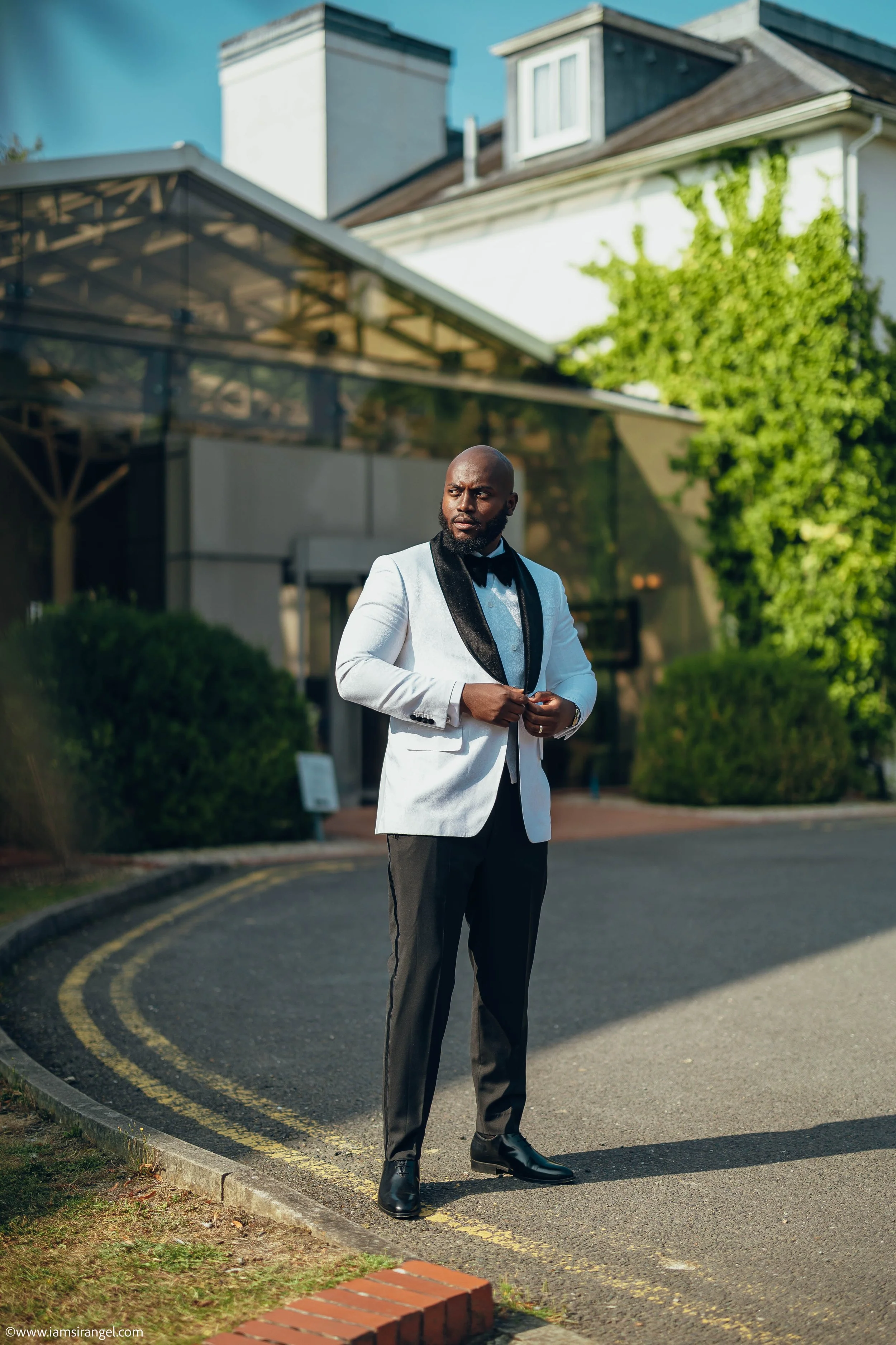 A man dressed in a white tuxedo jacket with a black lapel, black dress pants, and black dress shoes, standing outdoors near a building with green bushes and a tree, adjusting his coat.