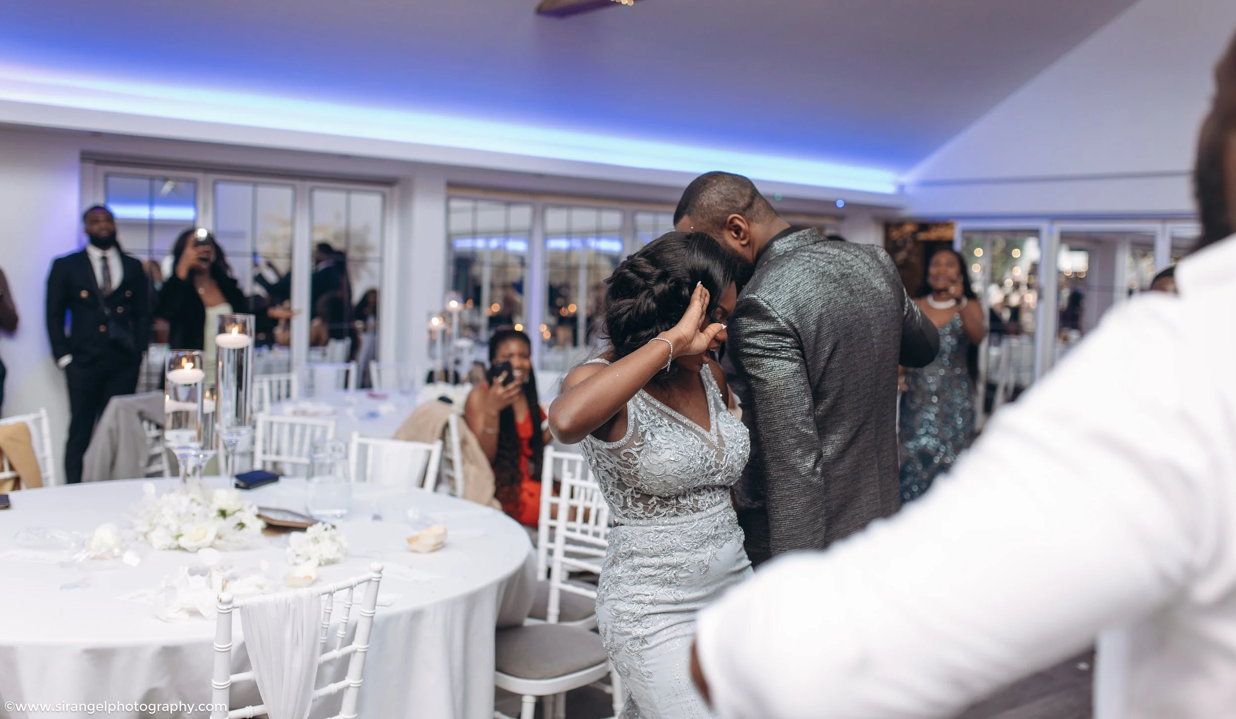 A couple dancing closely at a wedding reception, surrounded by friends and family in a decorated bright room.