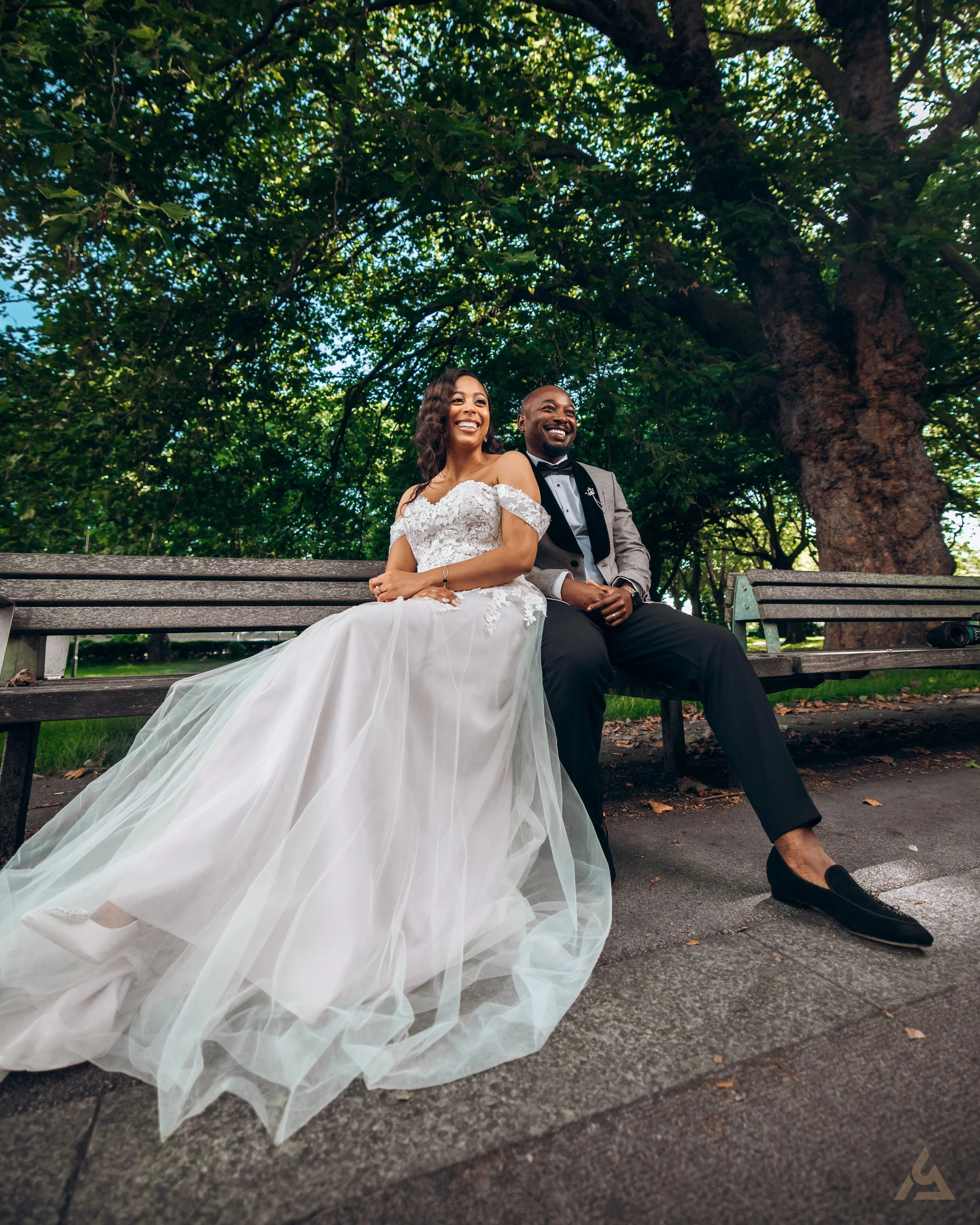 A bride and groom sitting on a park bench, smiling and enjoying each other's company, with lush green trees in the background.