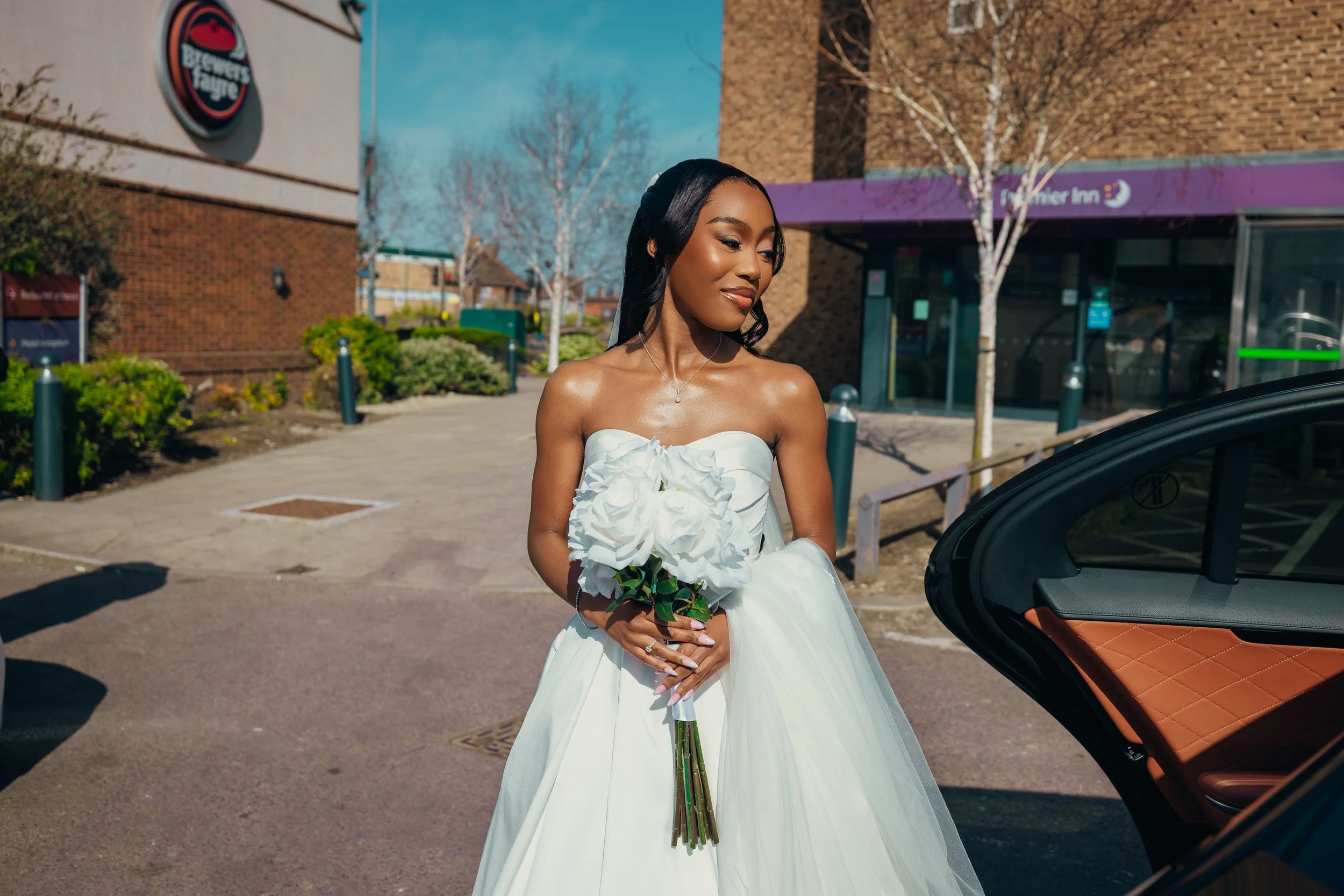Young woman in a white wedding dress holding a bouquet of white roses standing outside near a car, with a background of trees and buildings.