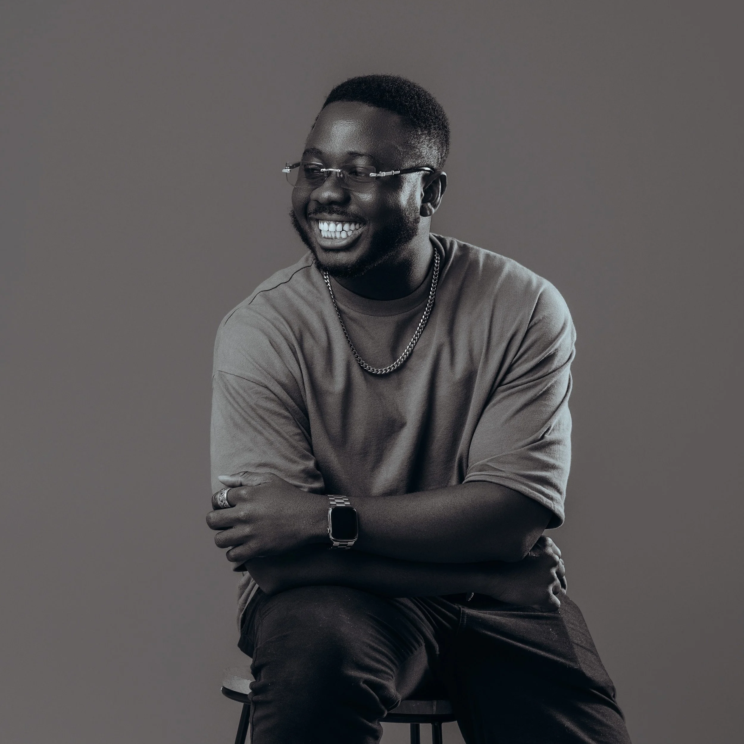 A monochrome photo of Sir Angel smiling, wearing a necklace and some sleek glasses. Sir Angel is a UK based Photographer who is loved by his clients. They say he serves good vibes only.