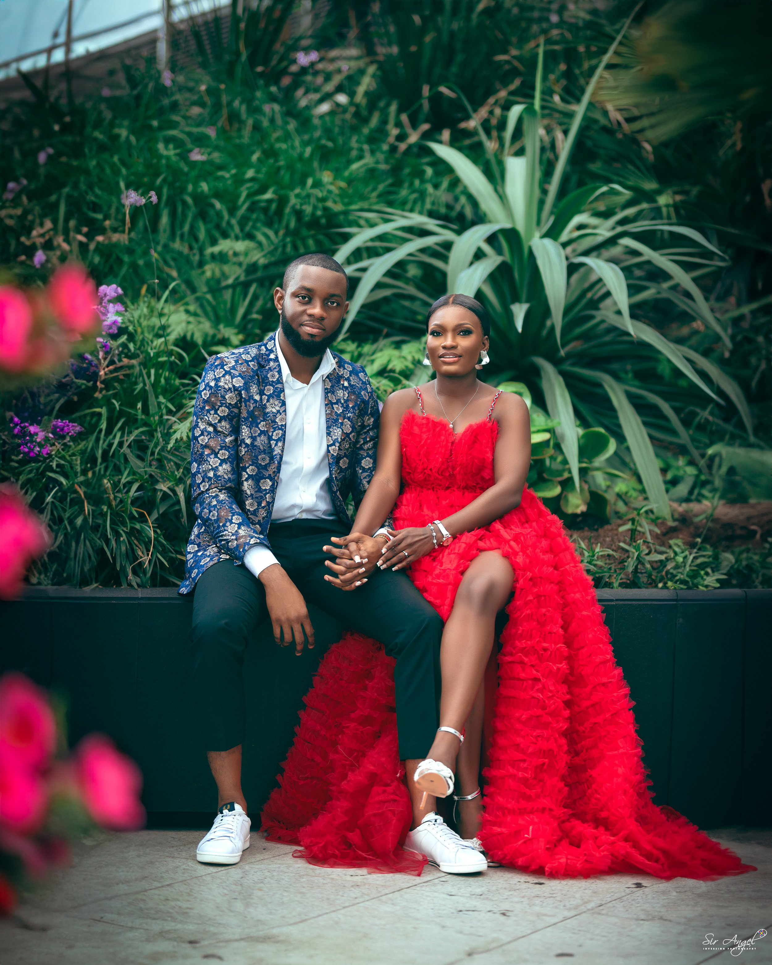 A couple sitting outdoors, the man dressed in a patterned blue blazer, white shirt, black pants, and white sneakers, and the woman wearing a vibrant red ruffled gown, white sneakers, and jewelry, with lush green plants in the background.