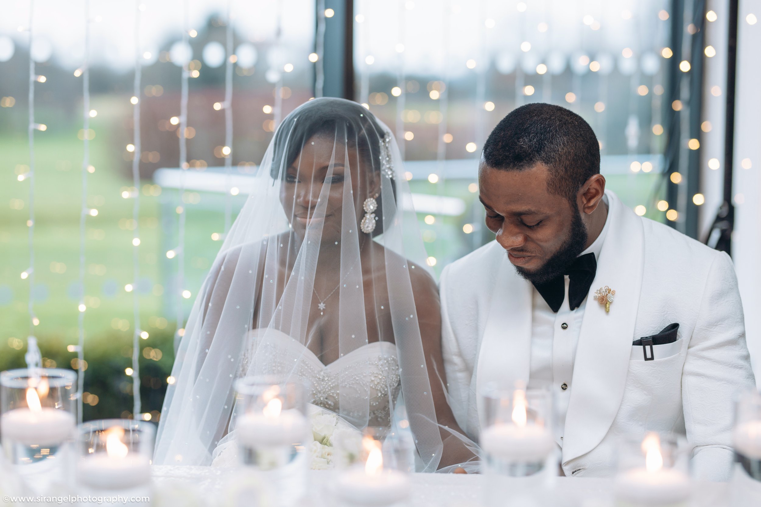 Bride and groom in wedding attire sitting at a table with lit candles, with fairy lights in the background.