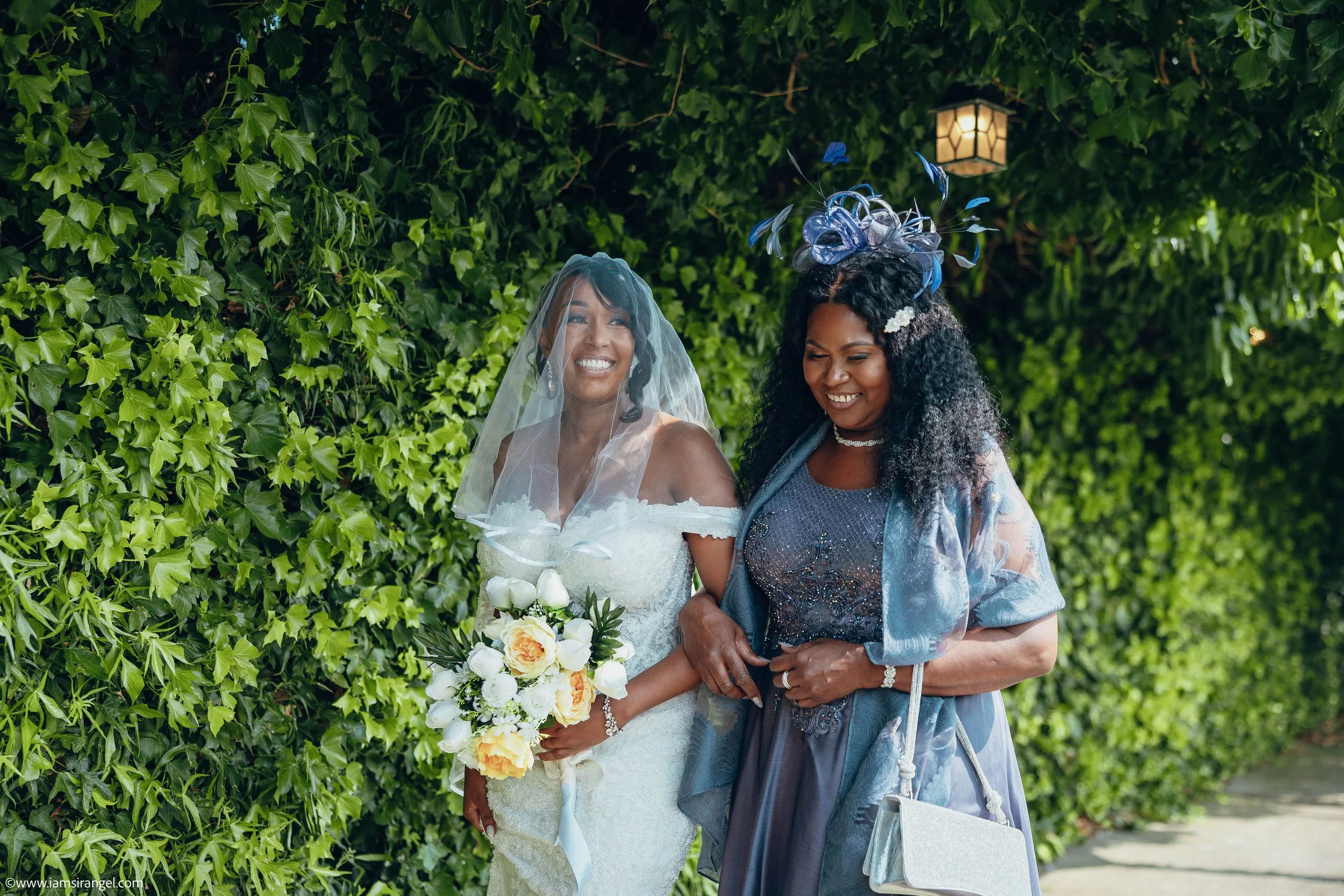 Two women, one in a wedding dress holding a bouquet, and the other in a fancy dress with a decorative headpiece, walk arm in arm outdoors near a green hedge, smiling and enjoying a special occasion.