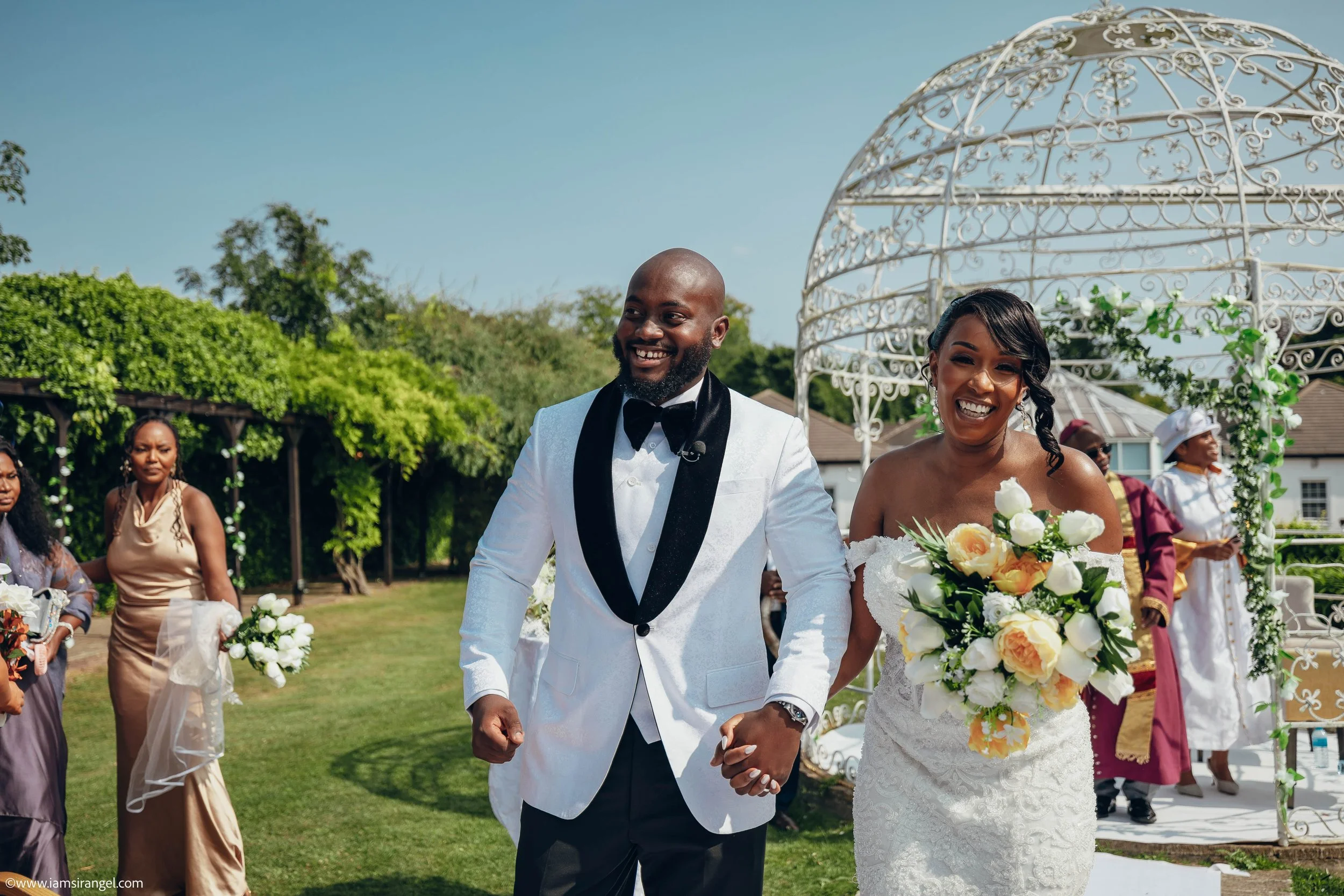 Bride and groom holding hands and smiling at their outdoor wedding ceremony, with bridal party and decorative arch in background.