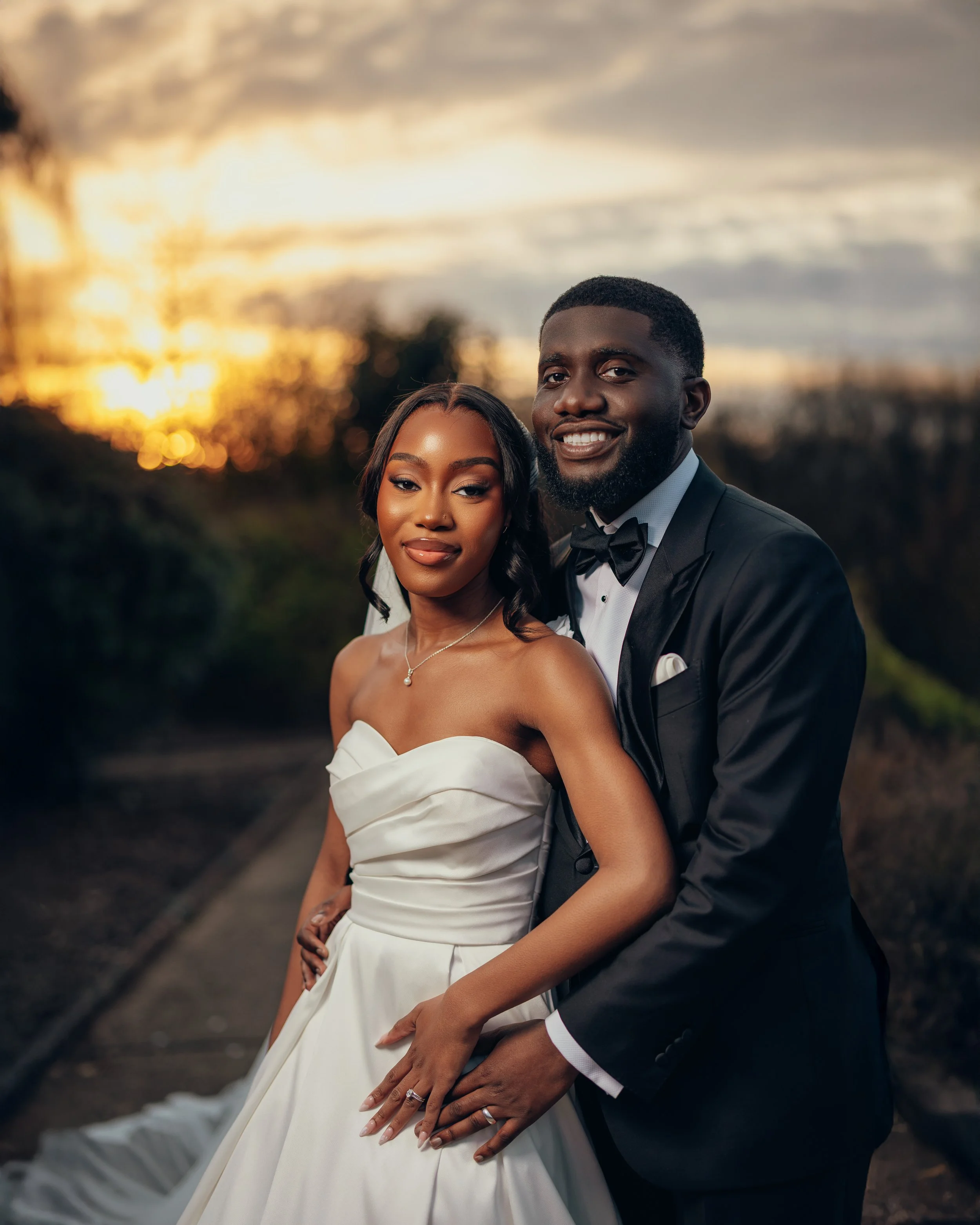 A newlywed couple, dressed in formal wedding attire, standing outdoors during sunset, smiling at the camera.