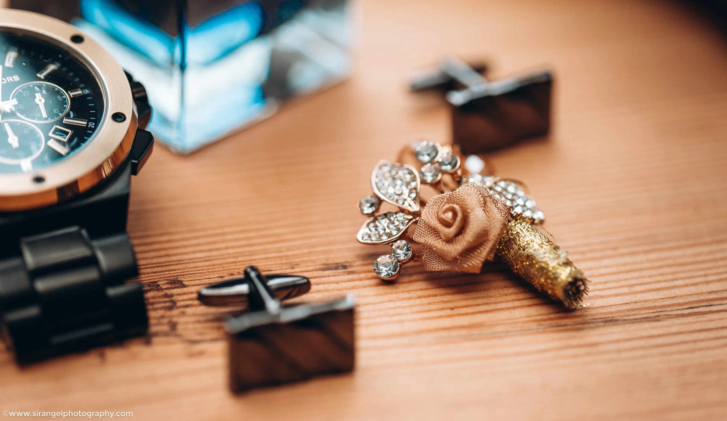 Close-up of a wristwatch, a pair of black earrings, a small square black object, and a decorative flower pin with rhinestones and gold glitter on a wooden surface.
