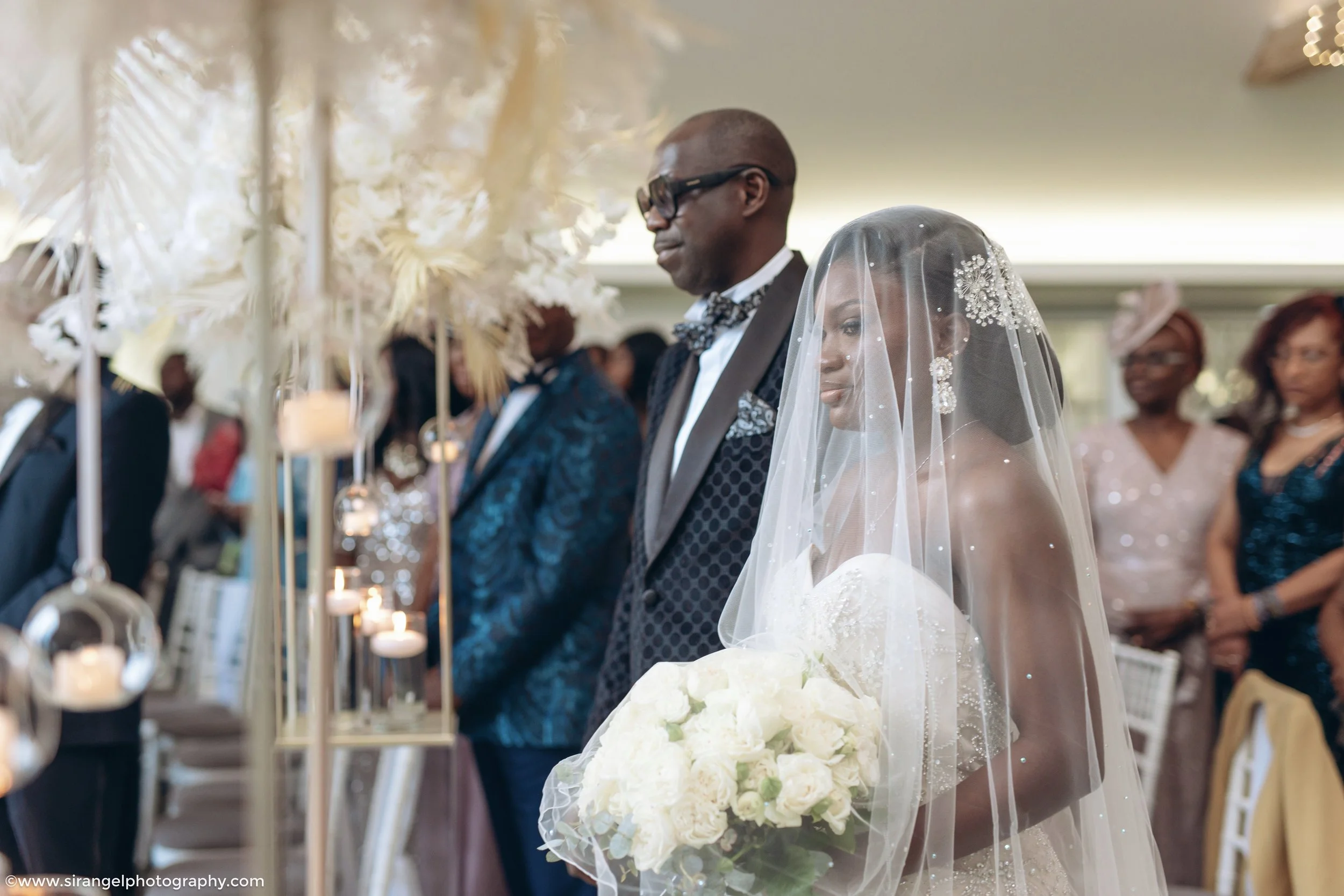 Bride holding a bouquet of white roses and wearing a veil, standing beside a man in a tuxedo, during a wedding ceremony with guests in the background.