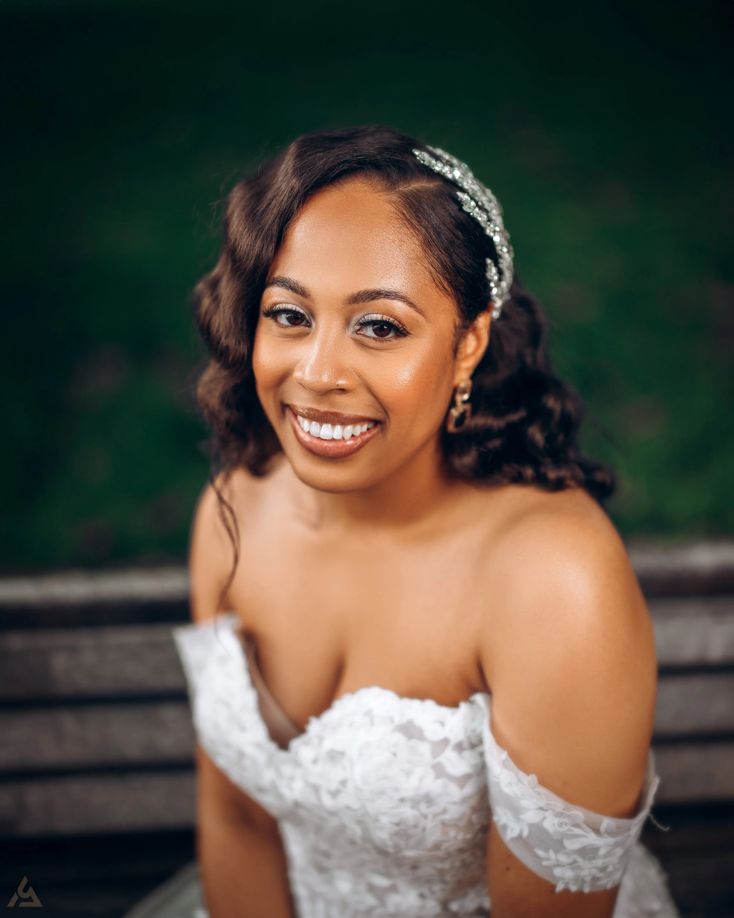 A smiling woman in a lace off-shoulder wedding dress, wearing earrings and a sparkly hair accessory, sitting on a park bench with a blurred green background.
