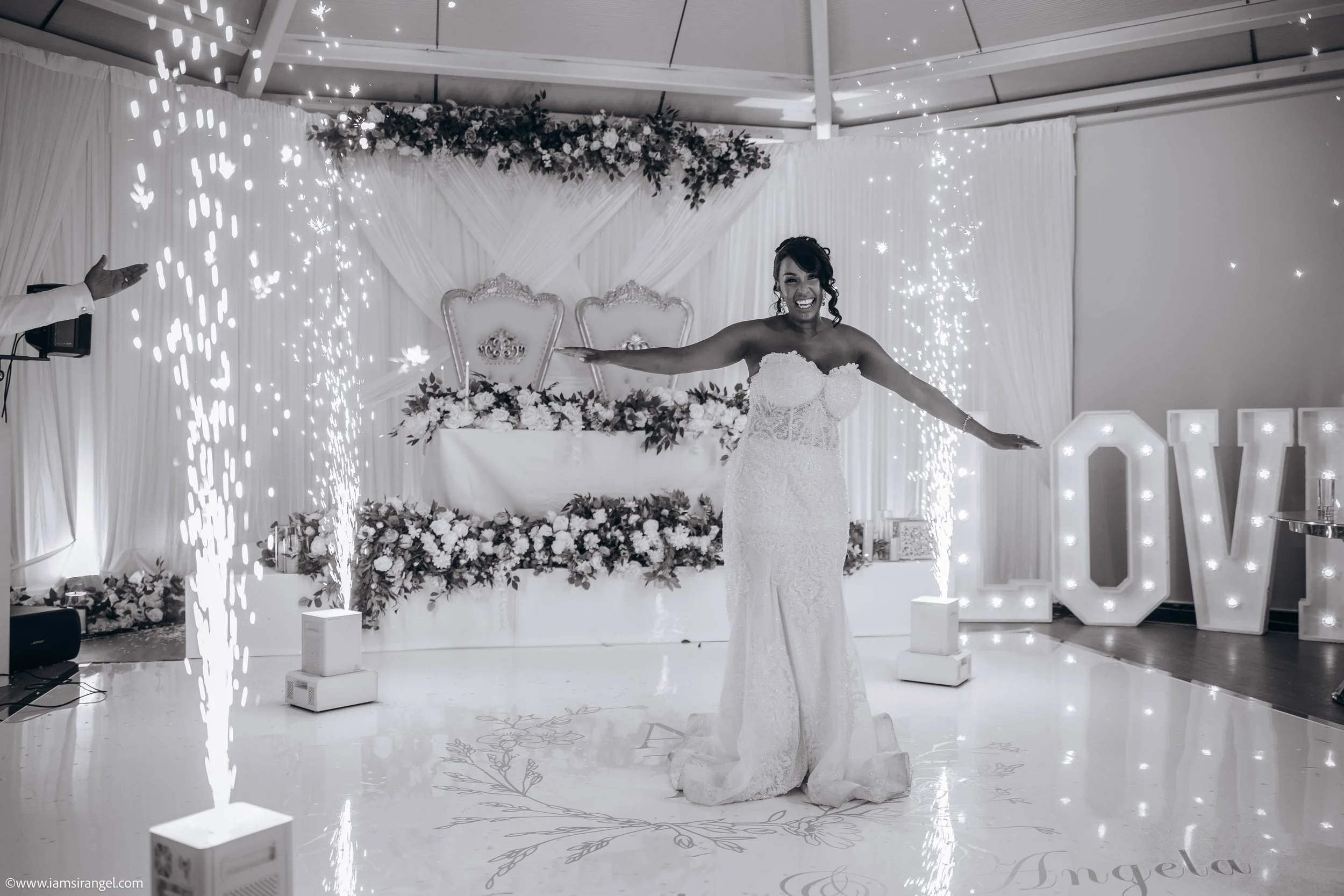 A bride in a wedding dress celebrating at her wedding reception with sparklers and illuminated letters spelling 'LOVE' in the background.