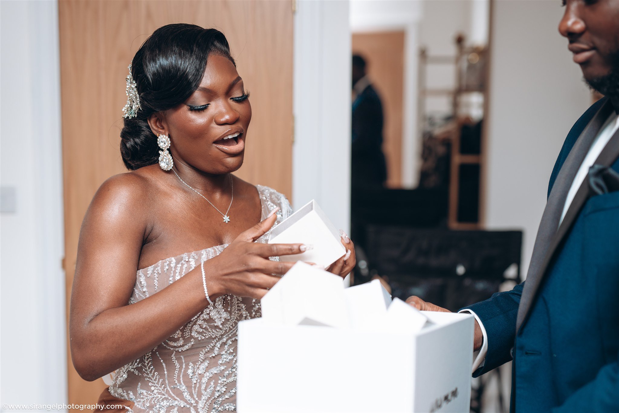 A woman in a wedding dress opening a gift box with a man in a suit watching at a wedding reception.