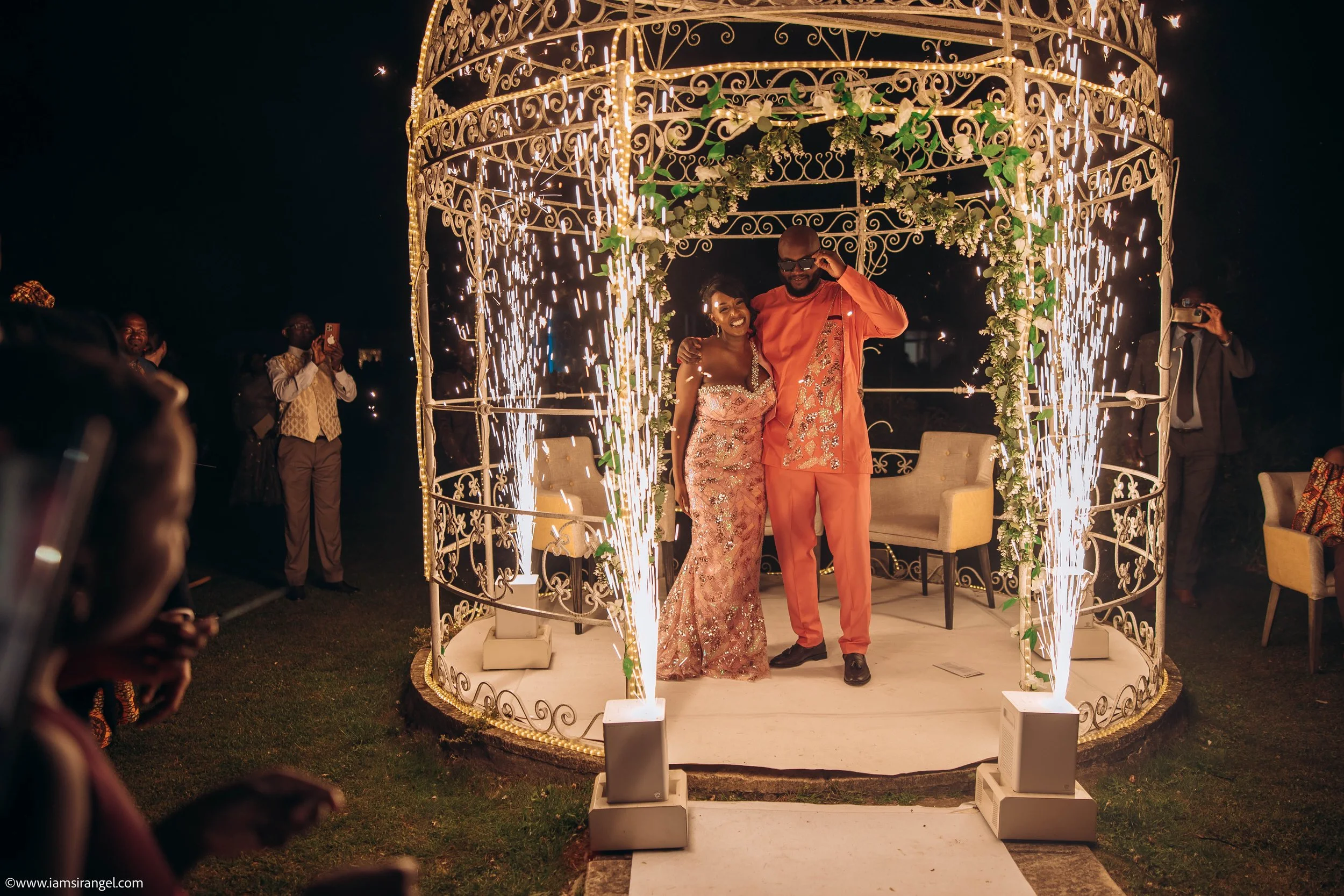 A couple is celebrating at a wedding or special event on a decorated stage with sparklers and string lights. The woman is wearing a pink, sparkly dress, and the man is dressed in a matching orange outfit with a patterned sash. They are smiling and em