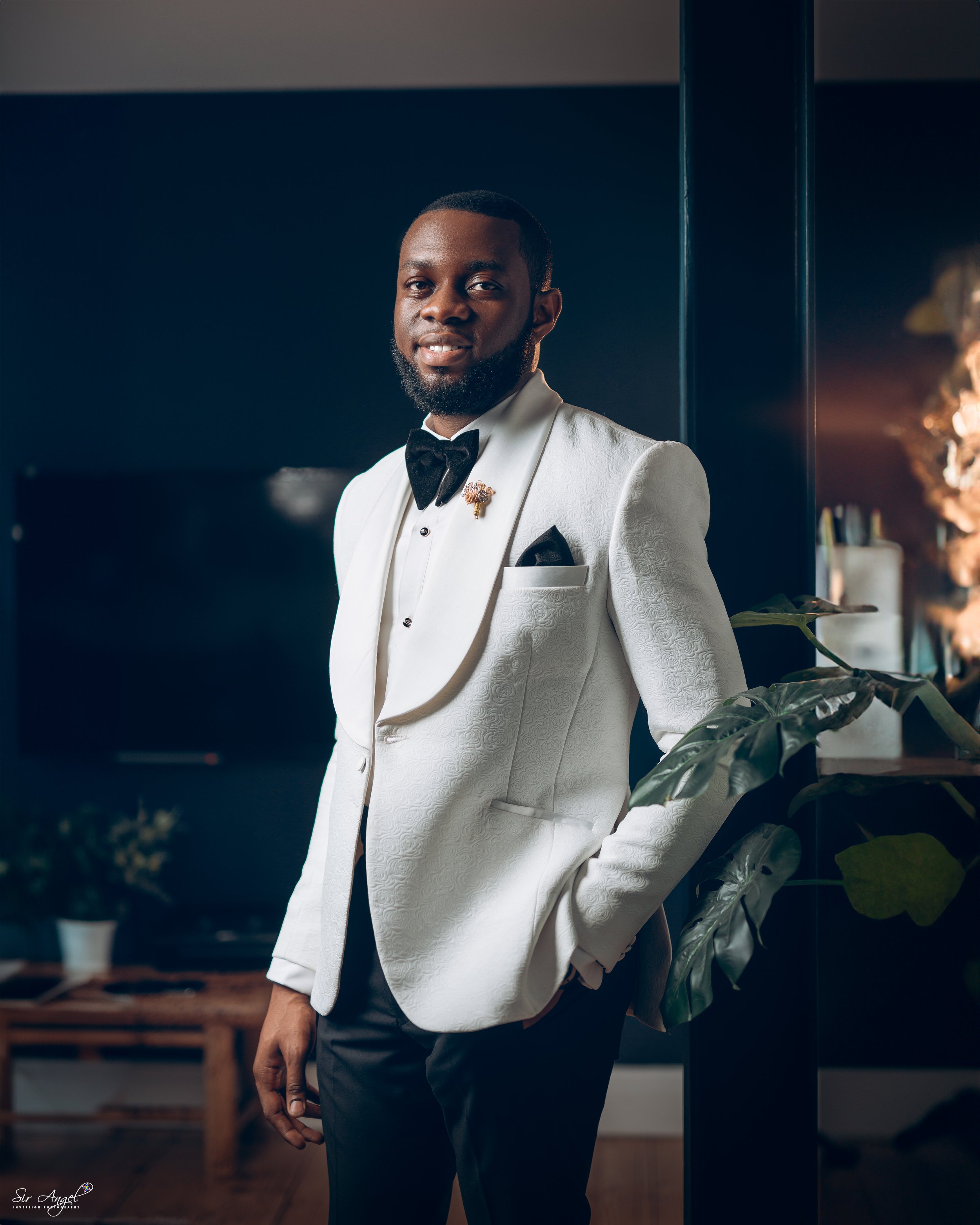 A man dressed in a white tuxedo with black bow tie and pocket square, standing indoors near a plant, smiling.