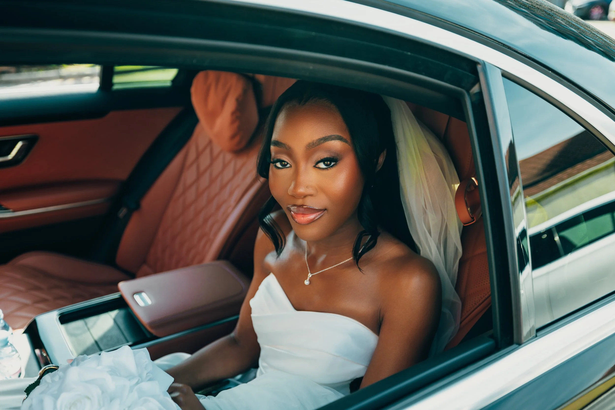 A bride sitting in the backseat of a car, wearing a white wedding dress and veil, looking at the camera with a slight smile.