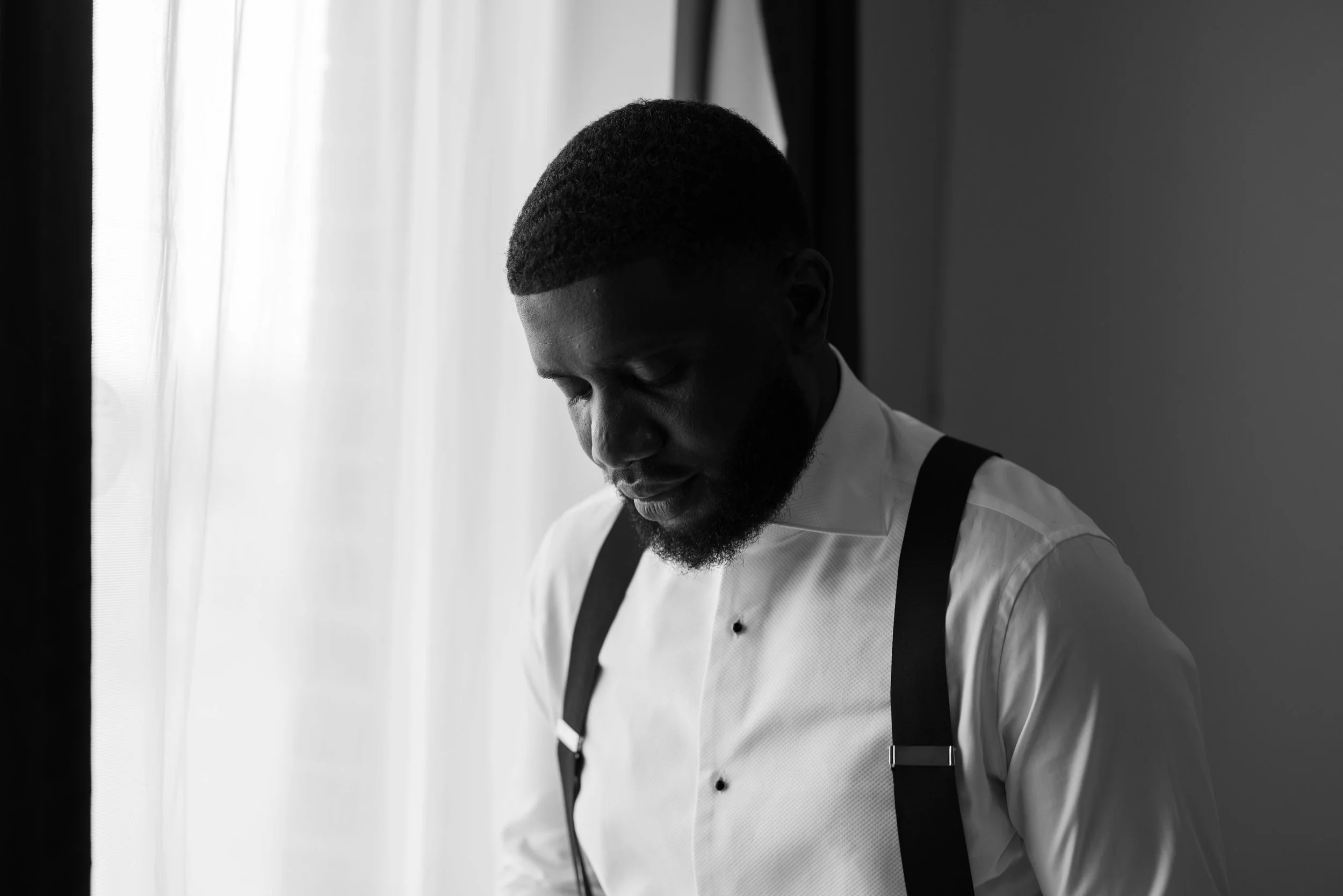 A black and white photo of a man with a beard in a white shirt and black suspenders, looking down, standing near a window with sheer curtains.