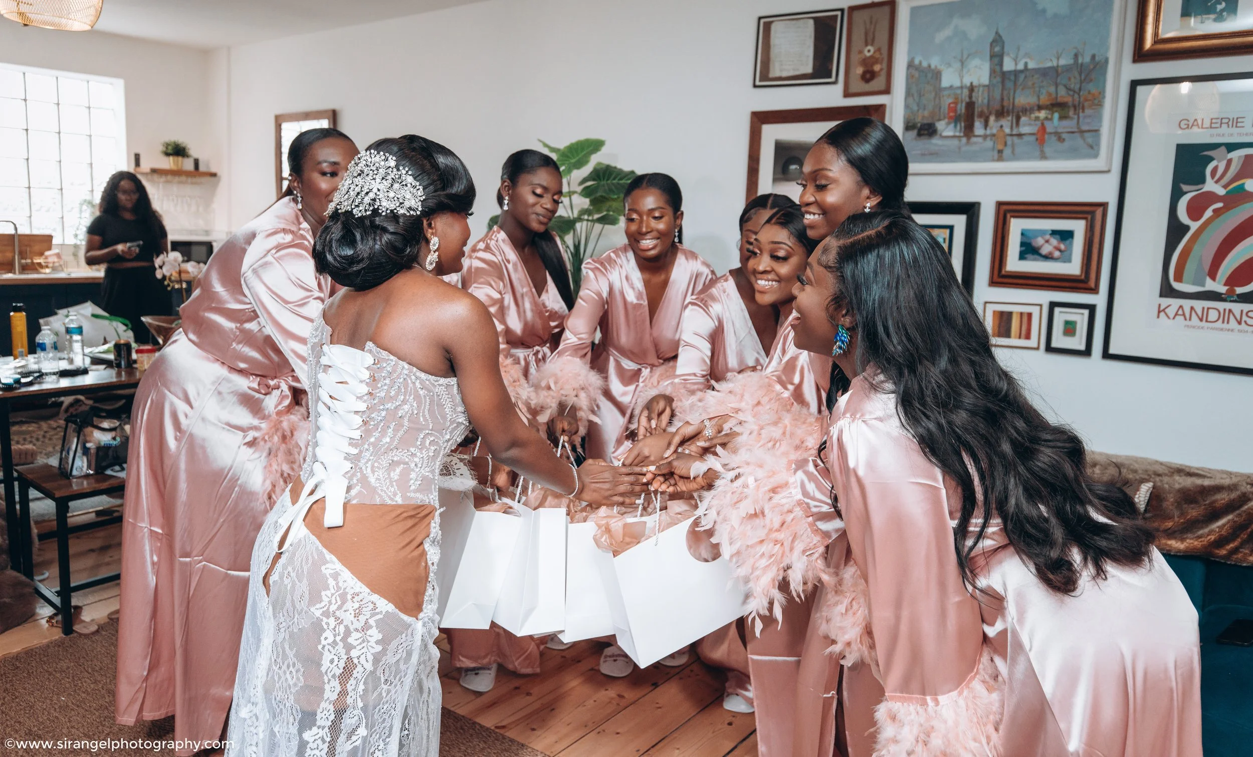 Bride and bridesmaids in pink satin robes celebrating together, exchanging gifts in a room decorated with framed artwork and certificates.
