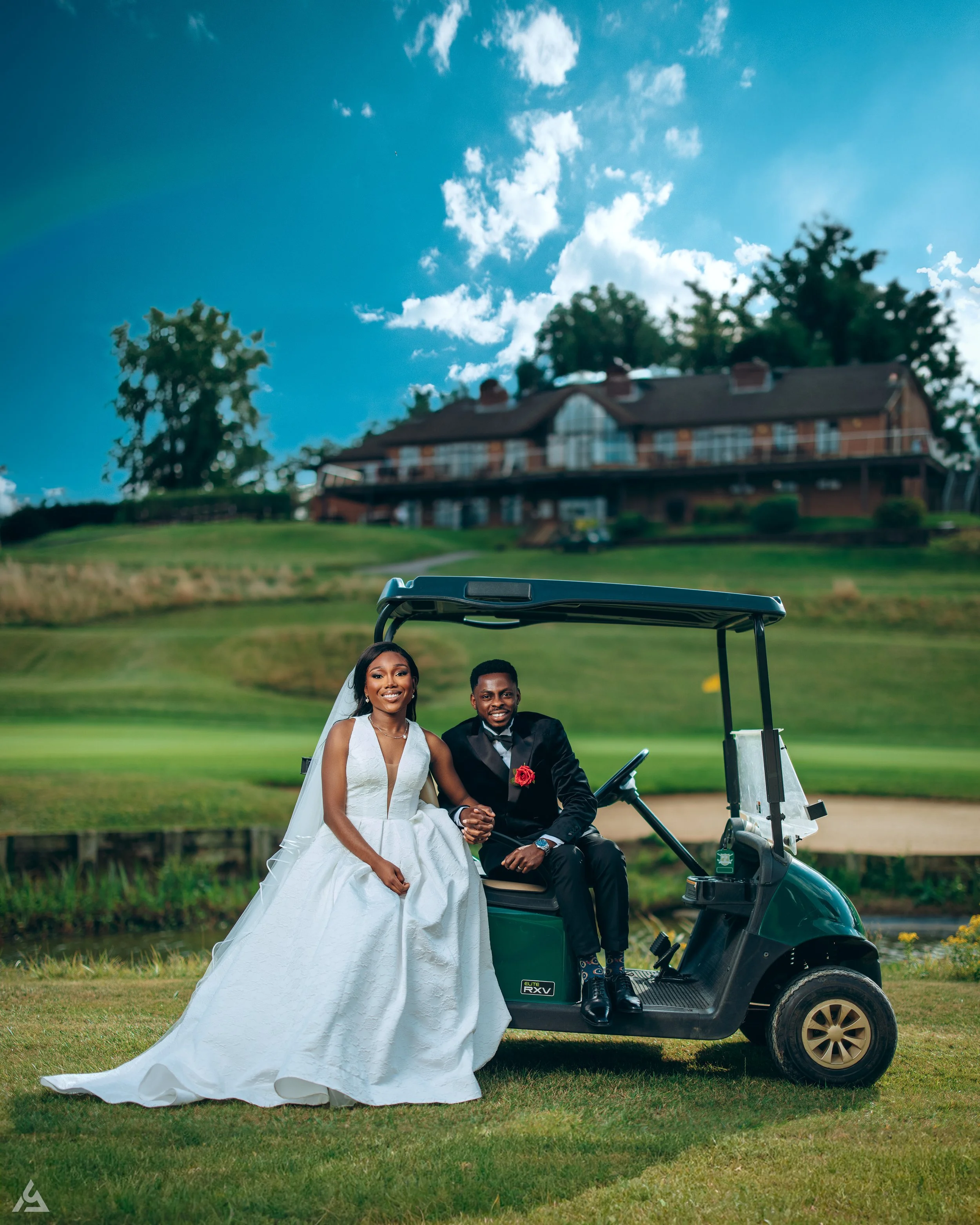 A bride and groom sitting on a golf cart on a golf course, with a large house in the background under a partly cloudy sky.