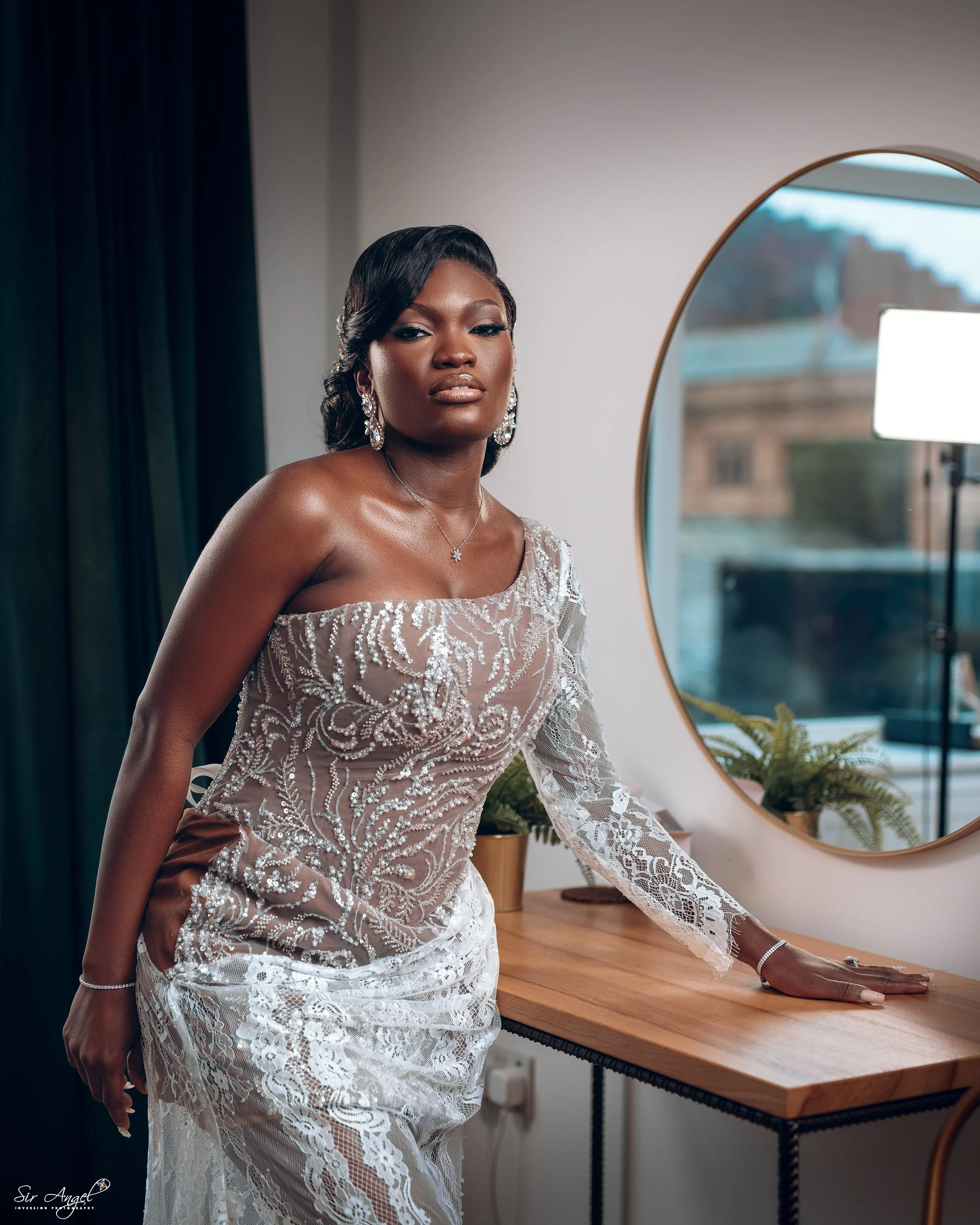 A woman dressed in a glamorous, detailed lace gown posing indoors near a wooden table and a round mirror.