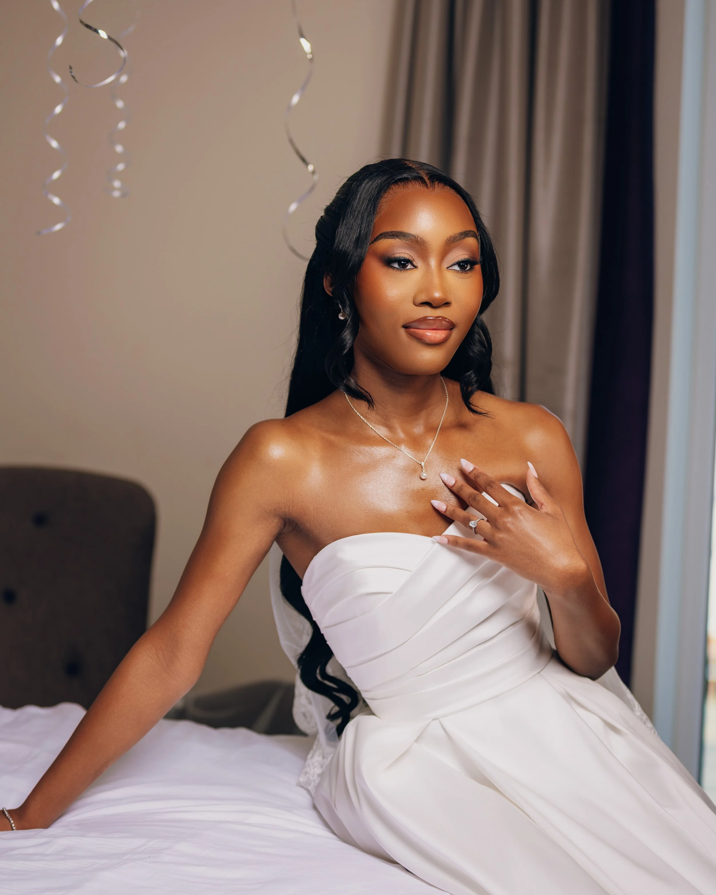 A woman in a wedding dress sitting on a bed, with her hand on her chest, wearing jewelry, with balloons and curtain in the background.
