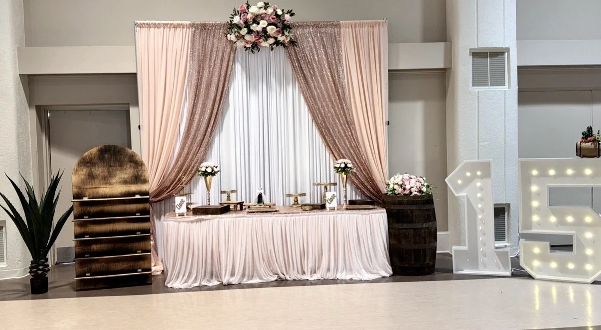 Decorative wedding or celebration setup with a pink pleated tablecloth, pink and white floral arrangements, and a backdrop with draped pink and glitter curtains. There are large light-up letters 'L' and 'E' on the right, and a wooden barrel with pink flowers on it. To the left, there is a dark green potted plant.