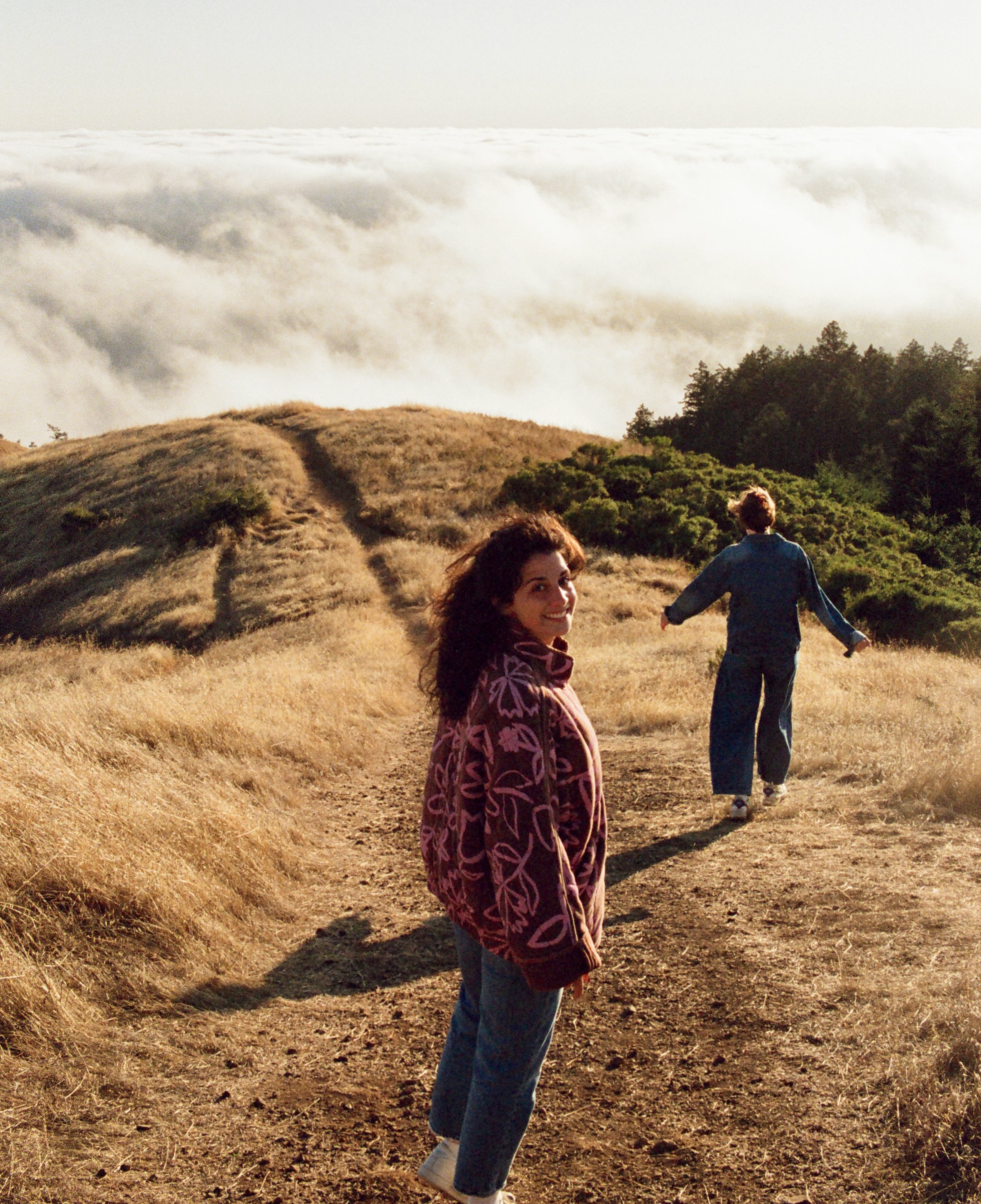Three people walking on a dirt trail through a grassy hillside with trees and clouds in the background in a natural outdoor setting.