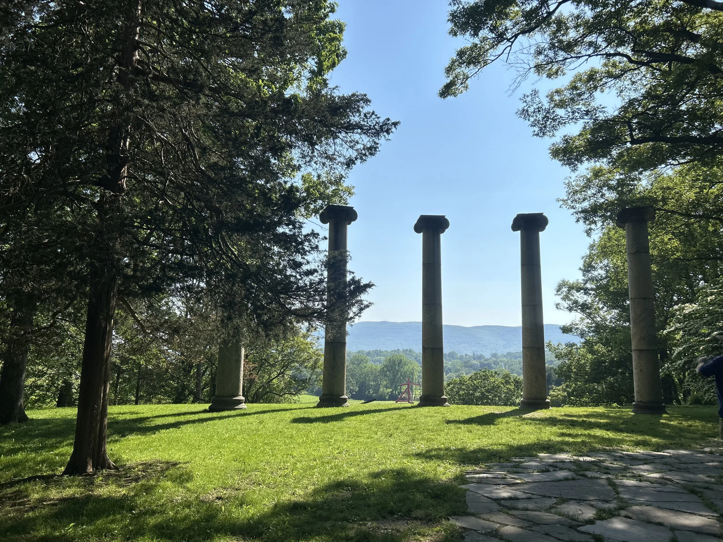 Ancient Greek columns standing in a grassy clearing, surrounded by tall trees with green leaves under a clear blue sky, mountain in the background.