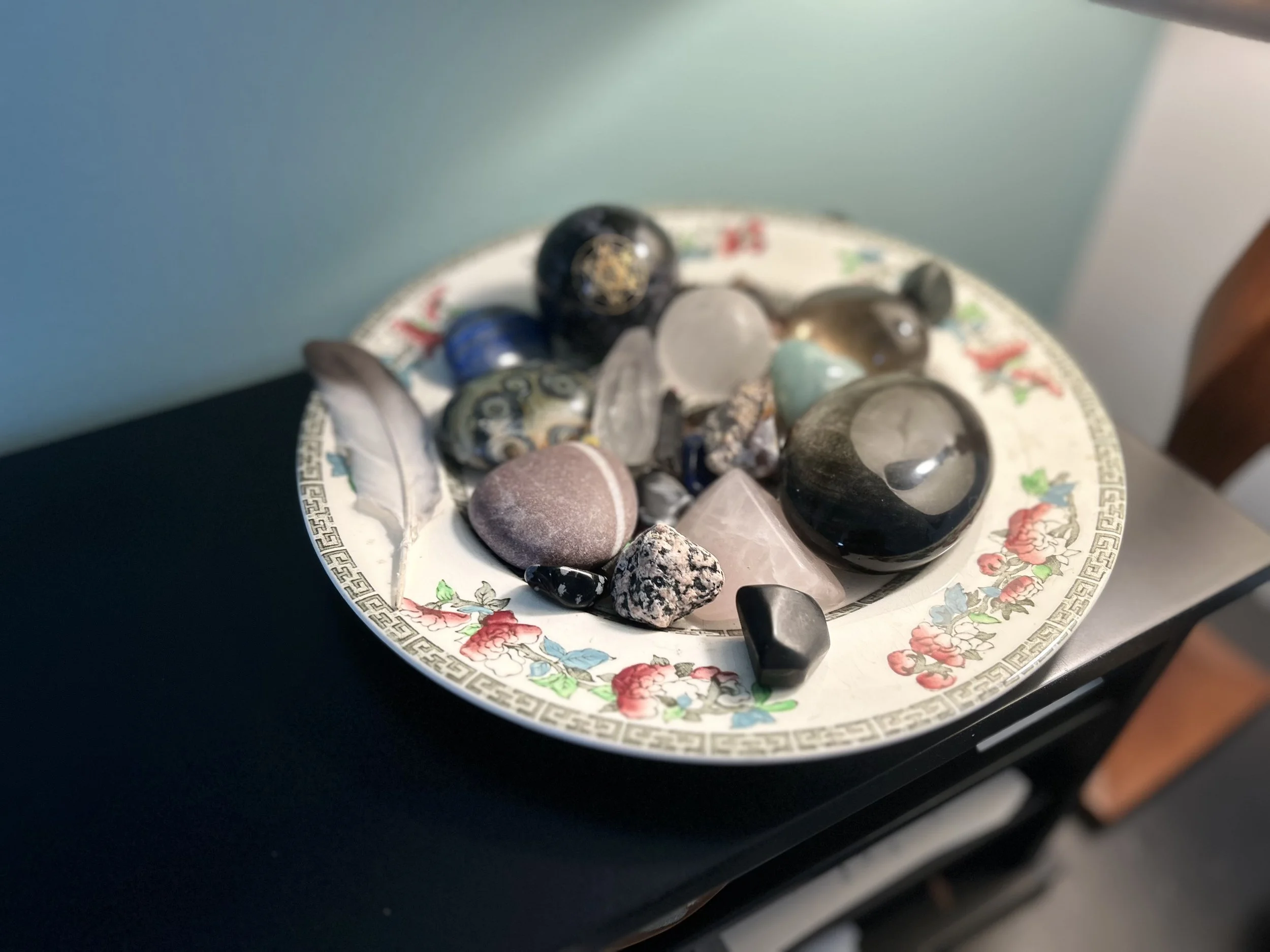 A decorative dish containing various polished stones and crystals of different shapes, sizes, and colors, with a patterned border and floral design in red, pink, blue, and green.